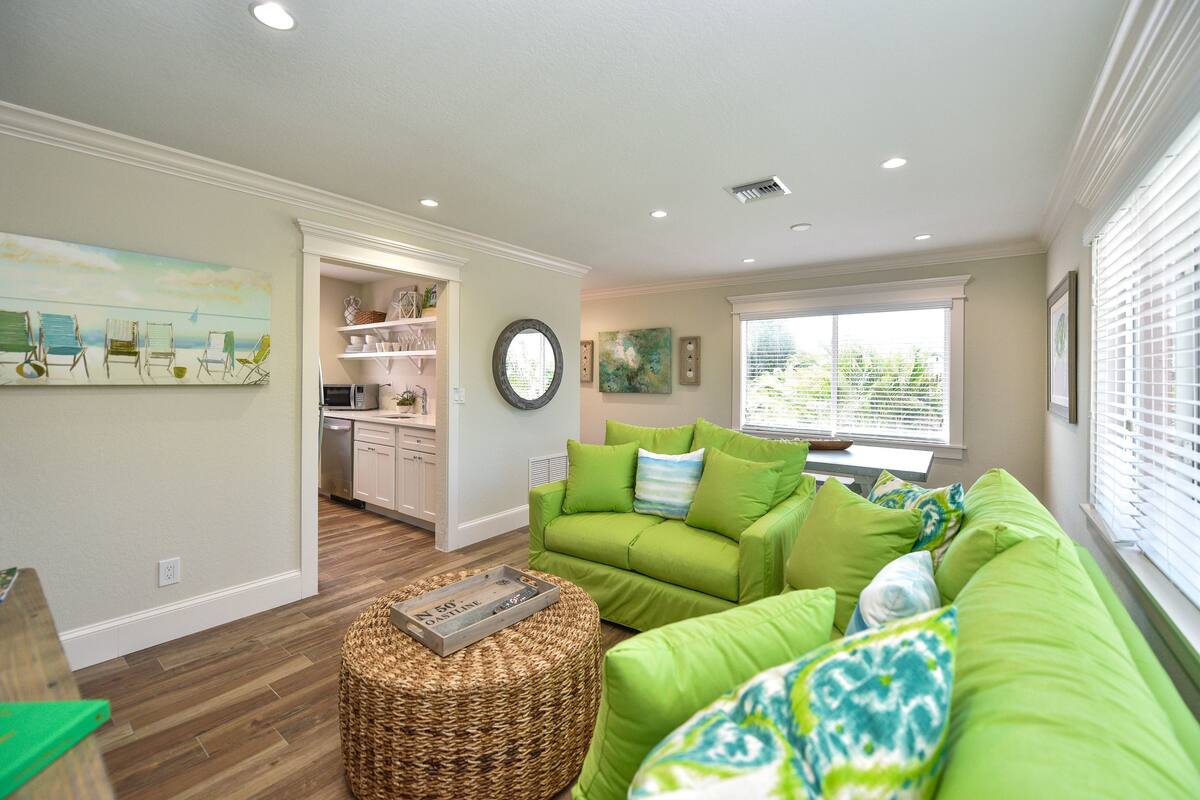 A living room with a green sofa, wicker ottoman, and a painting of beach chairs.