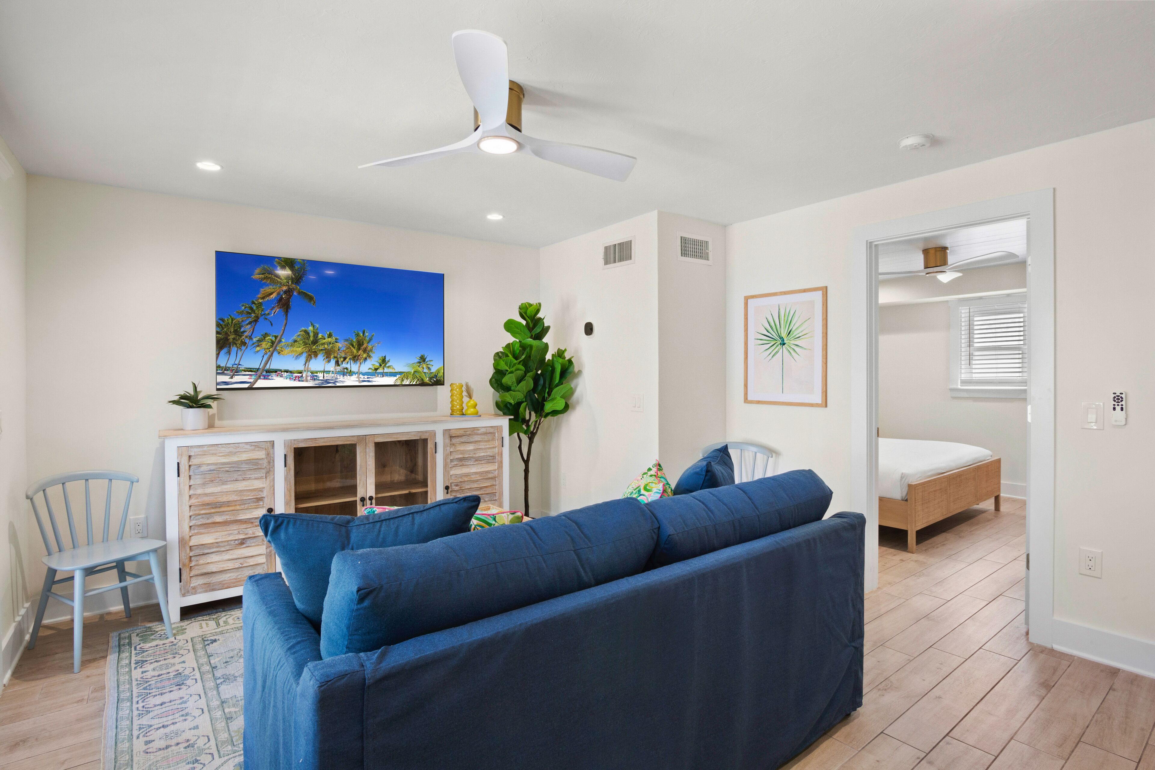 A living room with a blue sofa, a ceiling fan, a painting of palm trees, and a bed in the adjacent room.