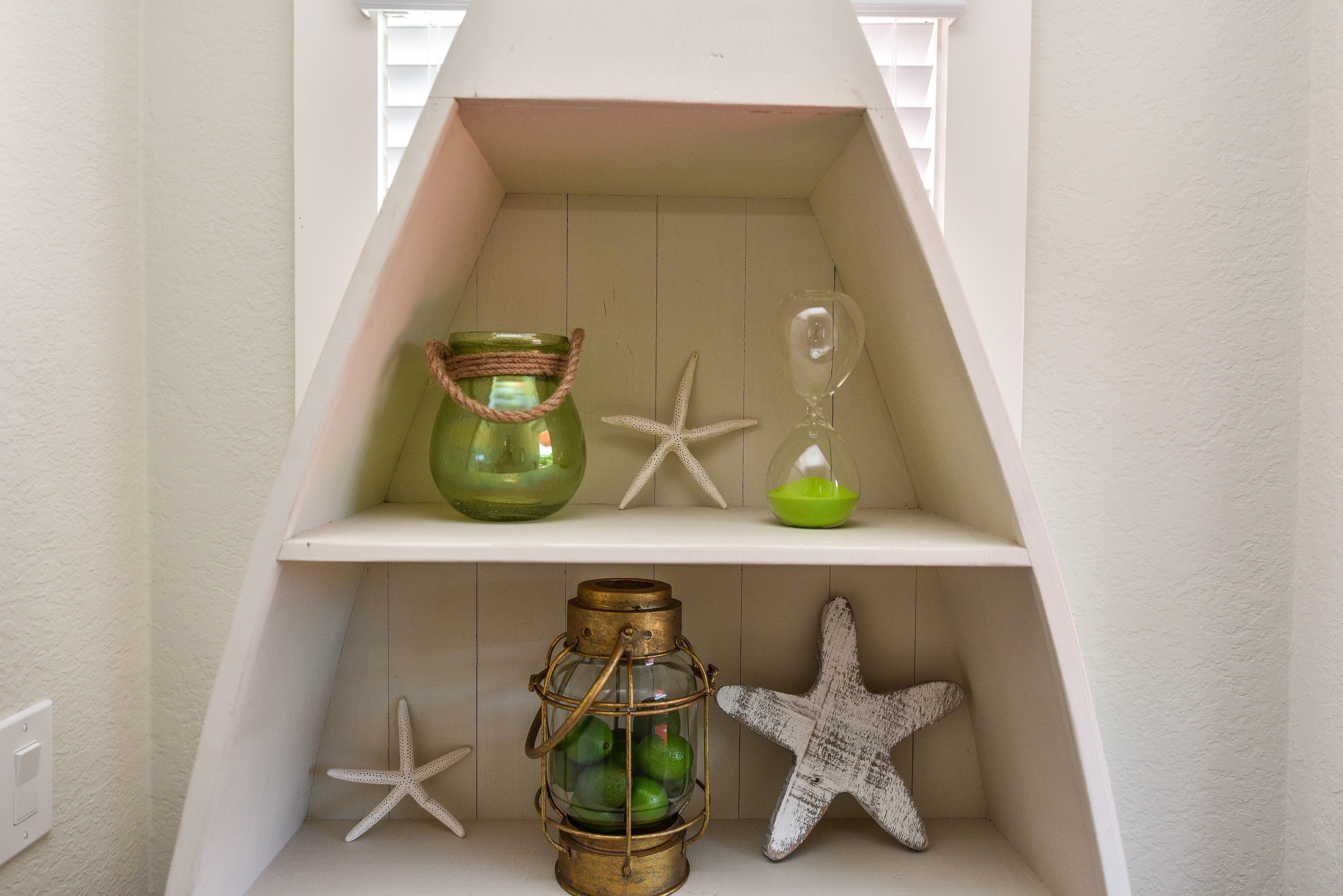 Un estante blanco con tres objetos decorativos: un frasco de vidrio verde con asa de cuerda, una ampolla de vidrio transparente y un cuenco de vidrio verde.