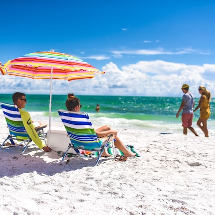 Two people relaxing under a striped beach umbrella on a sandy beach with the ocean in the background.
