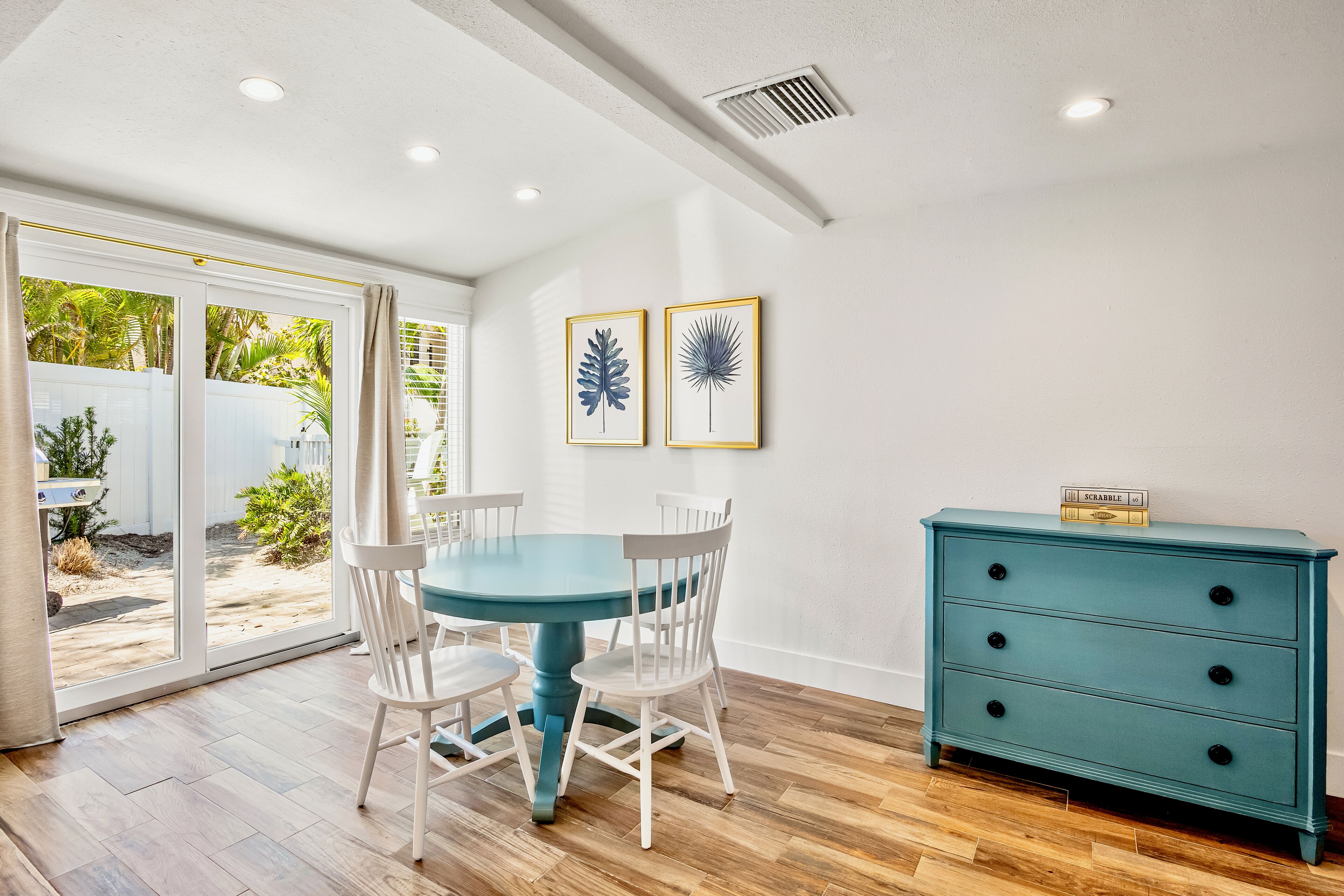 Un coin repas avec une table bleue et des chaises blanches, une commode turquoise et deux reproductions botaniques encadrées accrochées au mur.