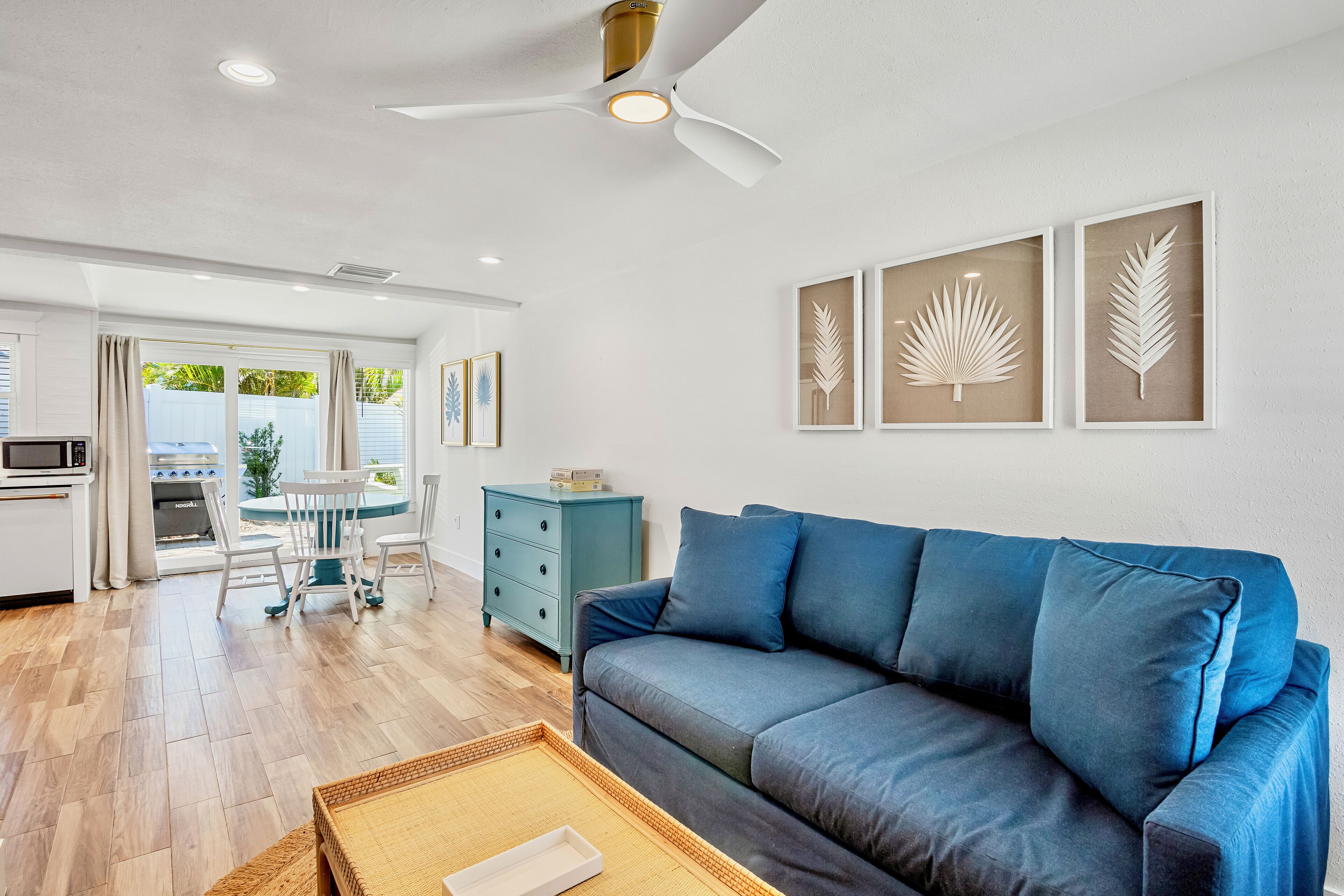 A modern living room with a blue sofa, a dining table with chairs, a teal dresser, and framed leaf artwork on the wall.