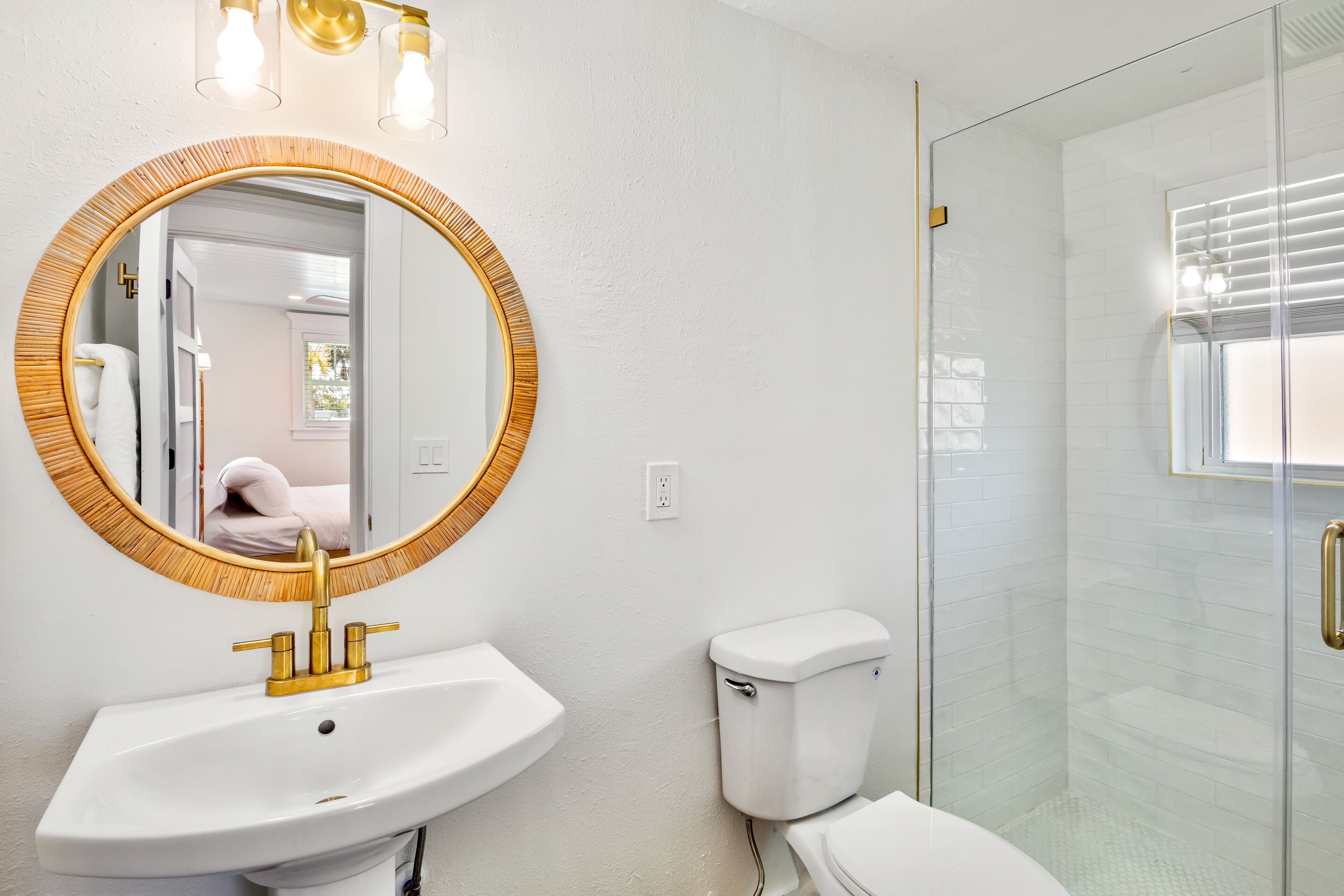 Een badkamer met een ronde spiegel, een wit wastafelmeubel met gouden details, een wit toilet en een douche met glazen wanden.