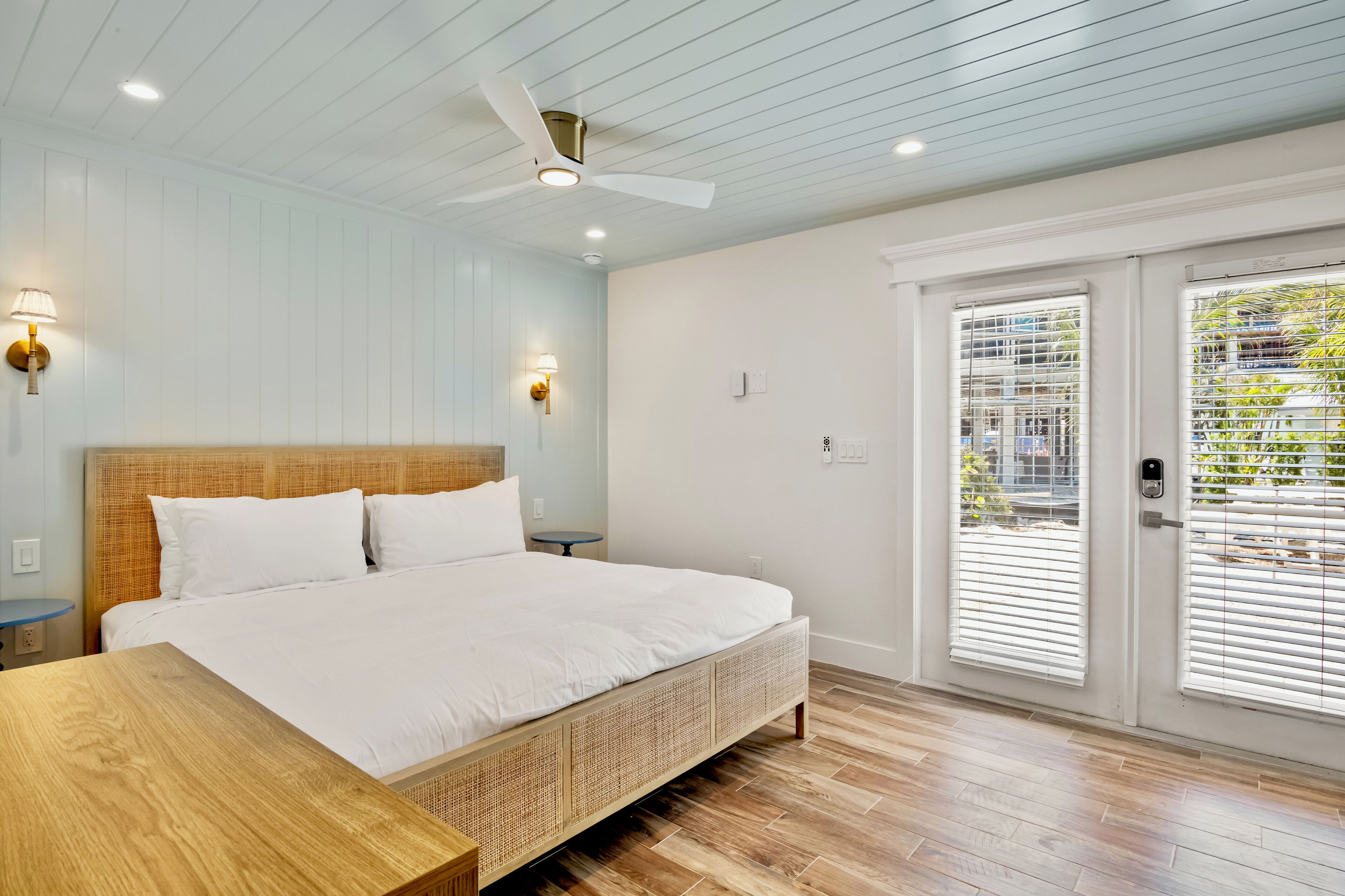 A bedroom with a bed, wooden headboard, ceiling fan, wall-mounted lights, and a view through a door with blinds.