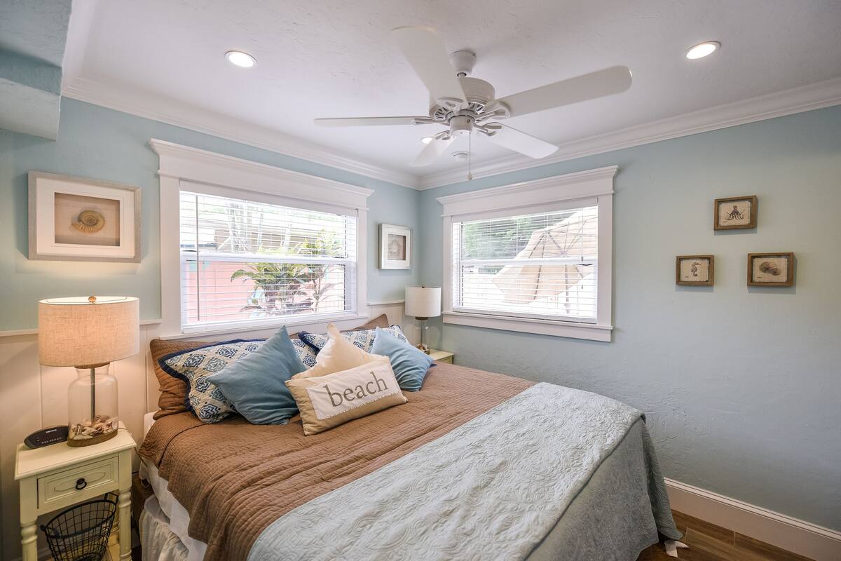 A bedroom with a bed, a nightstand, a lamp, a ceiling fan, and two windows with blinds.