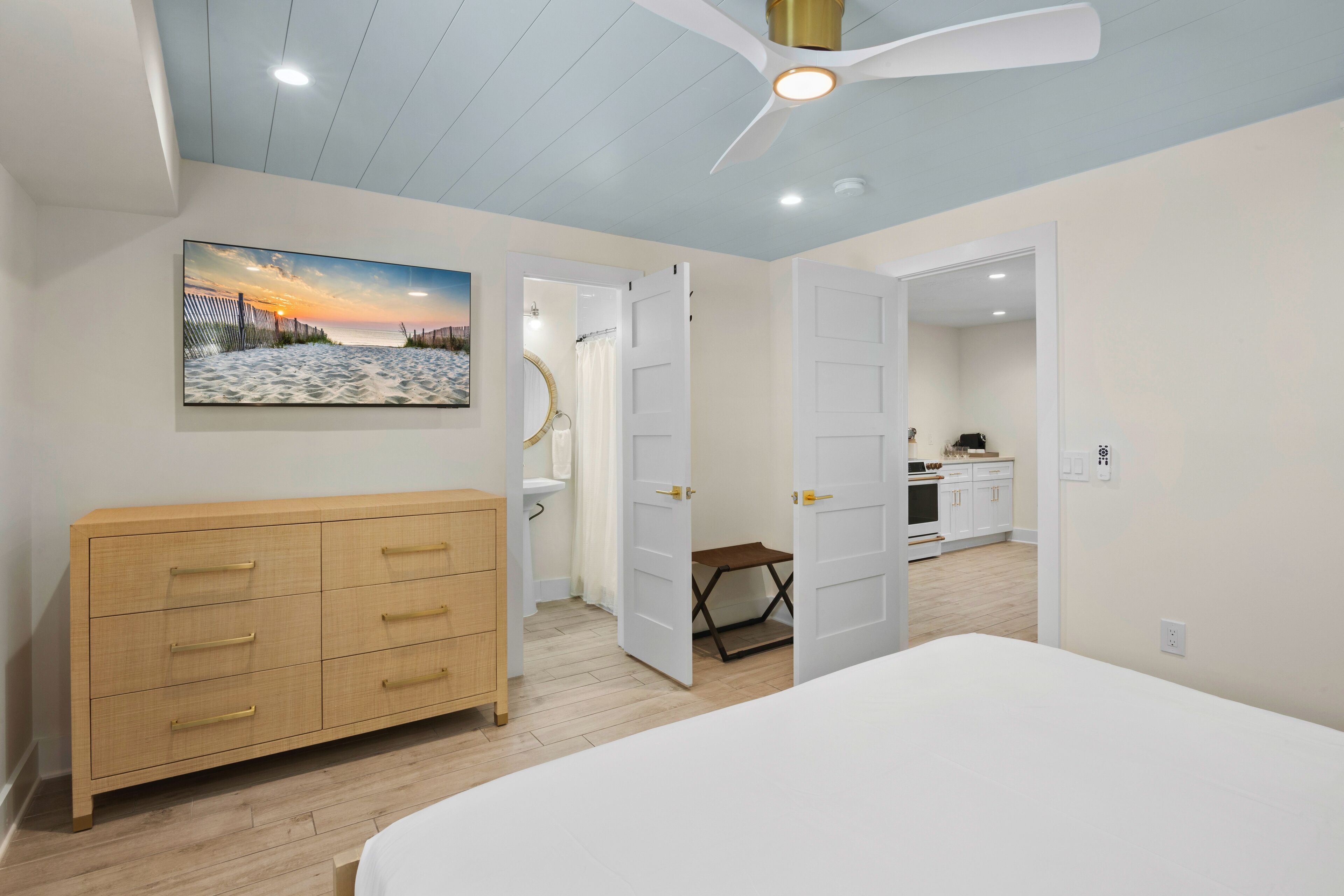 Une chambre à coucher comprenant un lit, une commode en bois, un ventilateur de plafond, un tableau représentant une scène de plage, et une porte ouverte donnant sur une salle de bain et une cuisine.