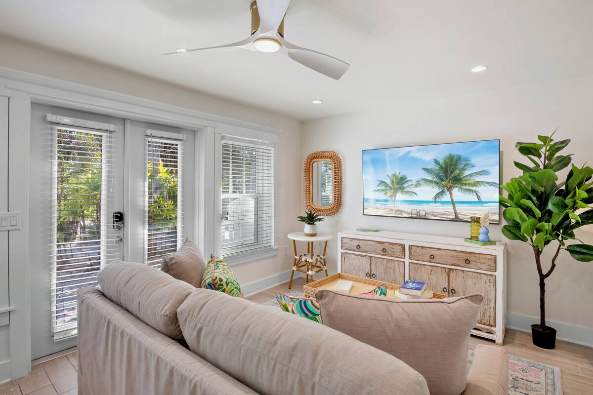 A living room with a beige sofa, a wooden console table, a ceiling fan, and a beach-themed painting.