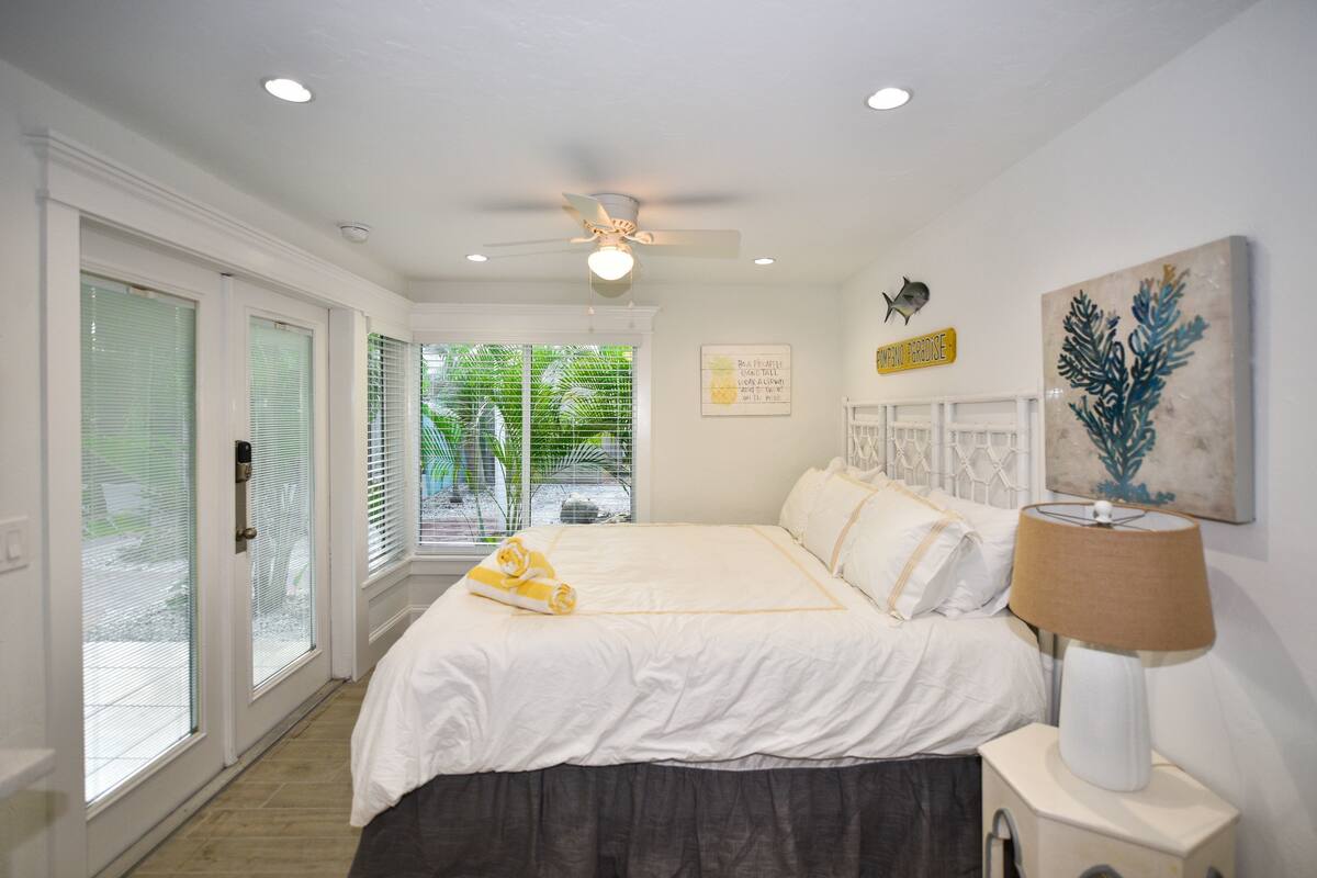 A bedroom with a large bed, a ceiling fan, a nightstand with a lamp, and a view of the outdoors through a sliding glass door.