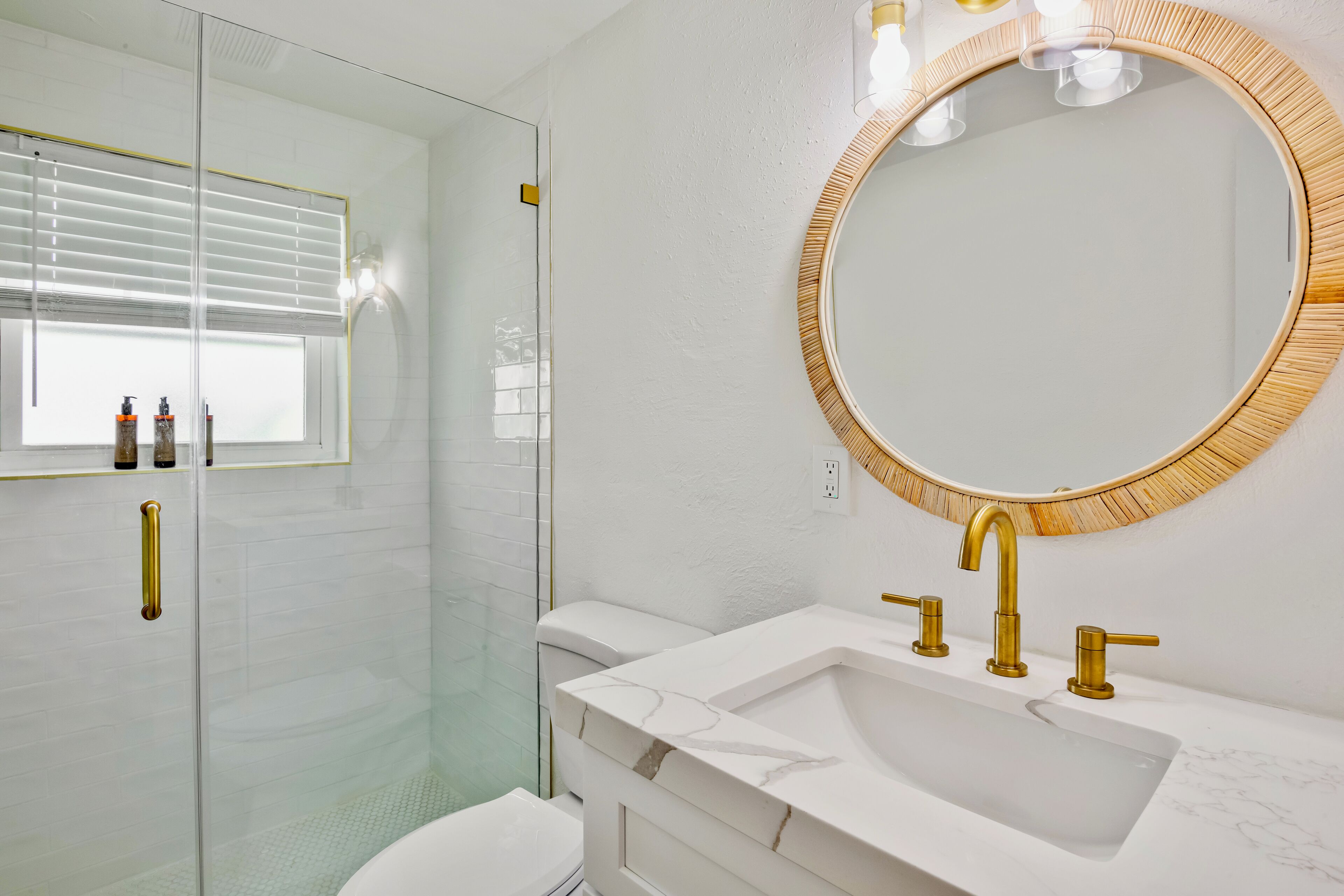 A modern bathroom with a glass shower enclosure, a white sink with gold fixtures, and a round mirror with a wooden frame.