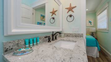 Un baño con encimera de granito, un lavabo blanco y un espejo grande. Se observan decoraciones de estrellas de mar en la pared y un sujetarrollos.