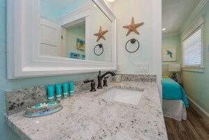 Un baño con encimera de granito, un lavabo blanco y un espejo grande. Se observan decoraciones de estrellas de mar en la pared y un sujetarrollos.