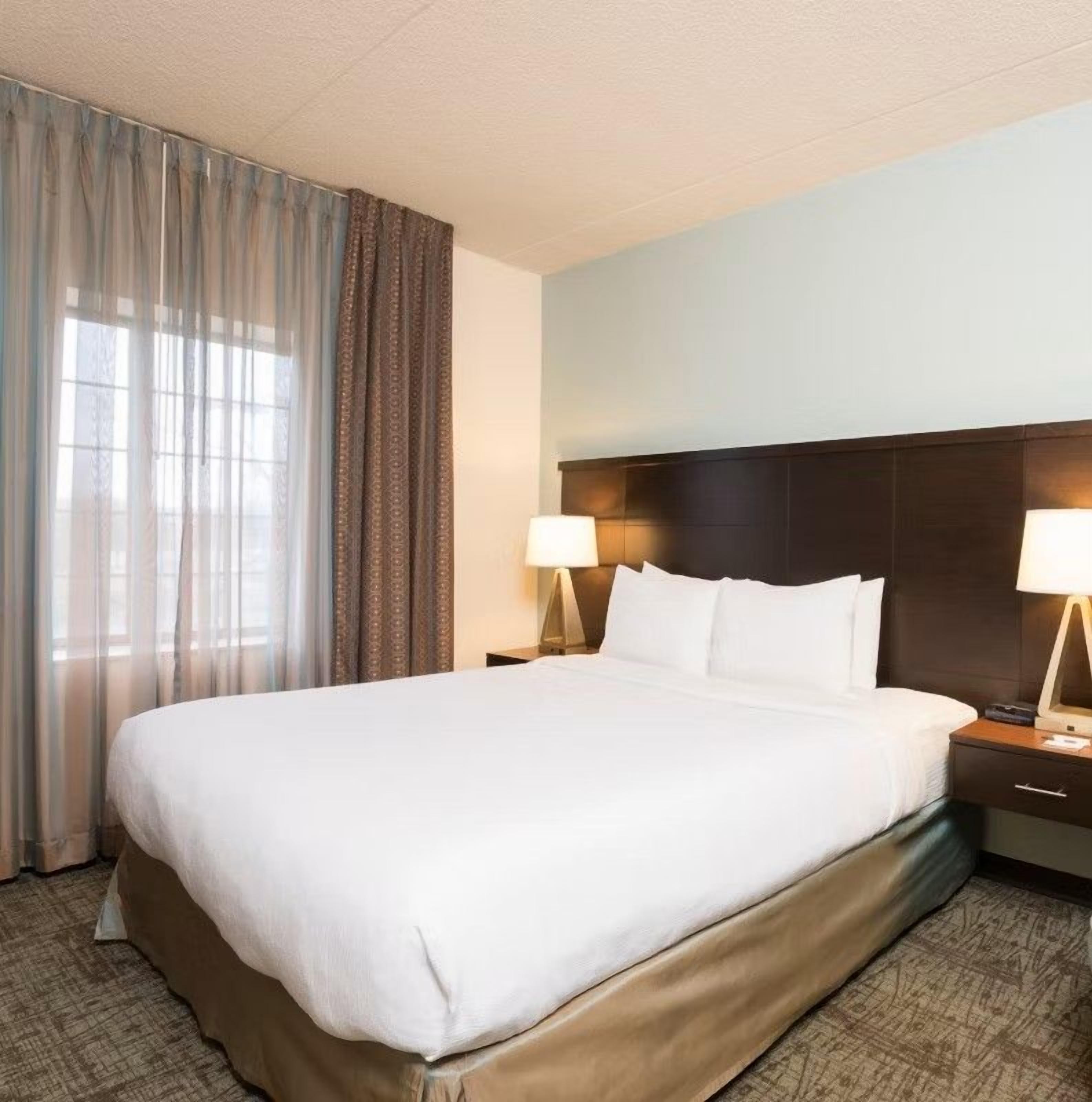 A hotel room with a large bed, two bedside tables, a headboard, and a window with curtains.