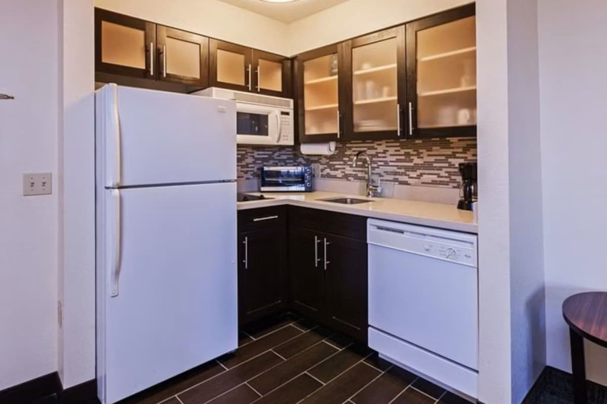 A compact kitchen with dark cabinets, a white refrigerator, a microwave, a dishwasher, and a tiled backsplash.