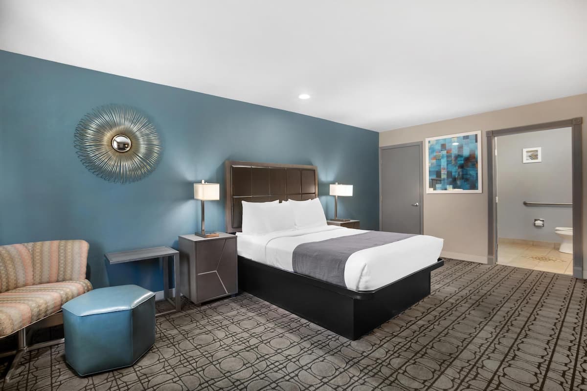 A hotel room with a bed, bedside tables, a chair, a wall art piece, and a bathroom door.
