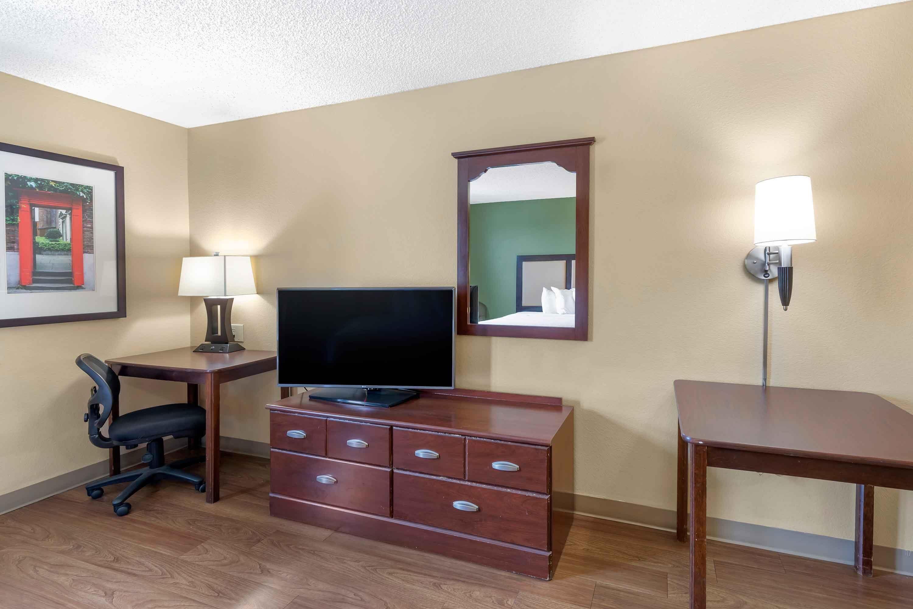Habitación de hotel con un escritorio de madera, televisor, cómoda con cajones, silla, lámpara, espejo y aplique en la pared.