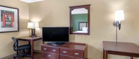 Habitación de hotel con un escritorio de madera, televisor, cómoda con cajones, silla, lámpara, espejo y aplique en la pared.