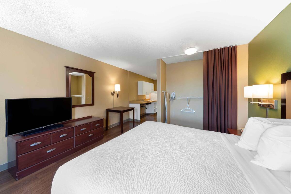 Une chambre d’hôtel avec un grand lit, un téléviseur posé sur une commode en bois, un bureau avec une chaise, un miroir et une salle de bain avec douche et toilettes.