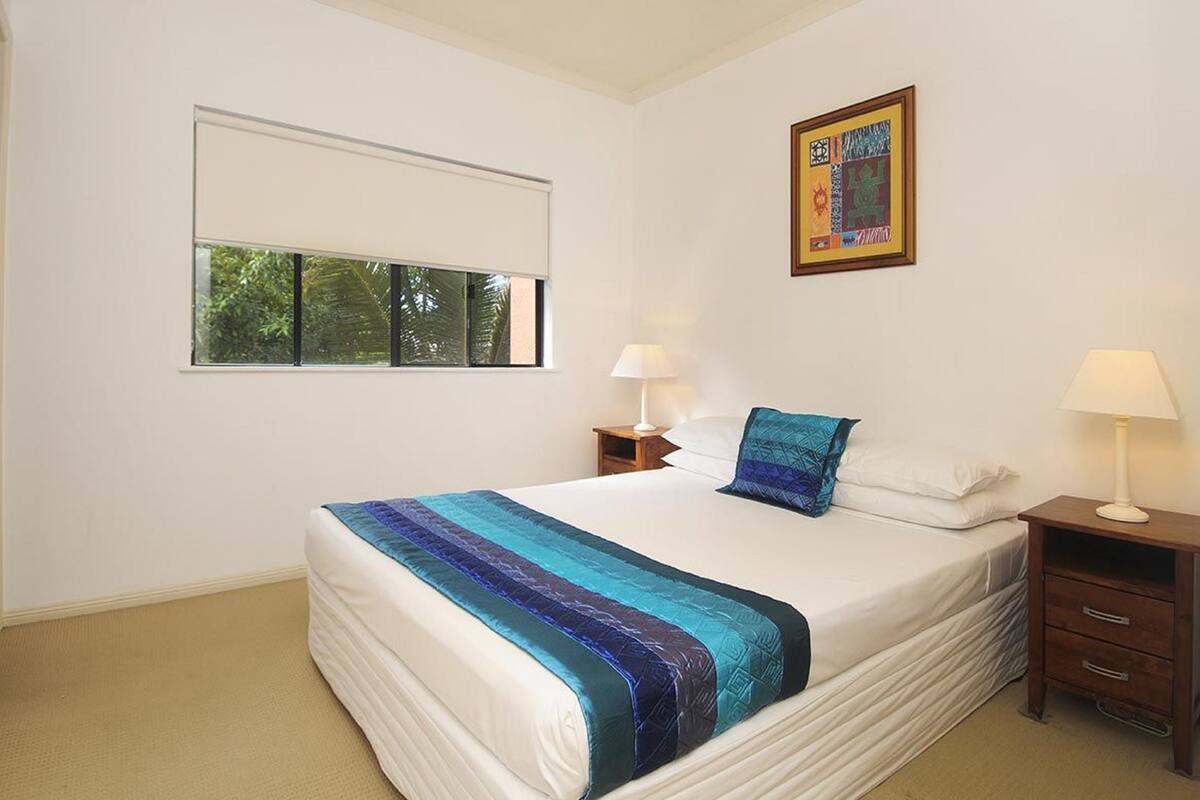 A bedroom with a large bed, two bedside tables with lamps, a window with blinds, and a framed artwork on the wall.