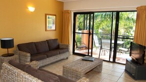 A living room with a sofa, a coffee table, a TV, and a balcony with chairs and a table. - Nautilus Holiday Apartments (Port Douglas)