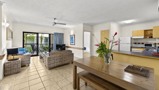 A modern living area with wicker furniture, a wooden dining table, and a kitchen with white appliances. - Nautilus Holiday Apartments (Port Douglas)