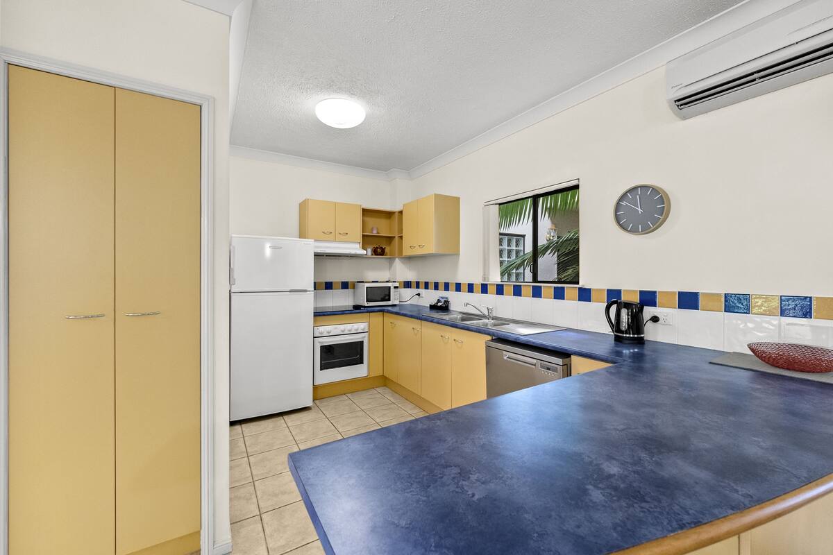 A kitchen with a blue countertop, white refrigerator, yellow cabinets, and a clock on the wall.