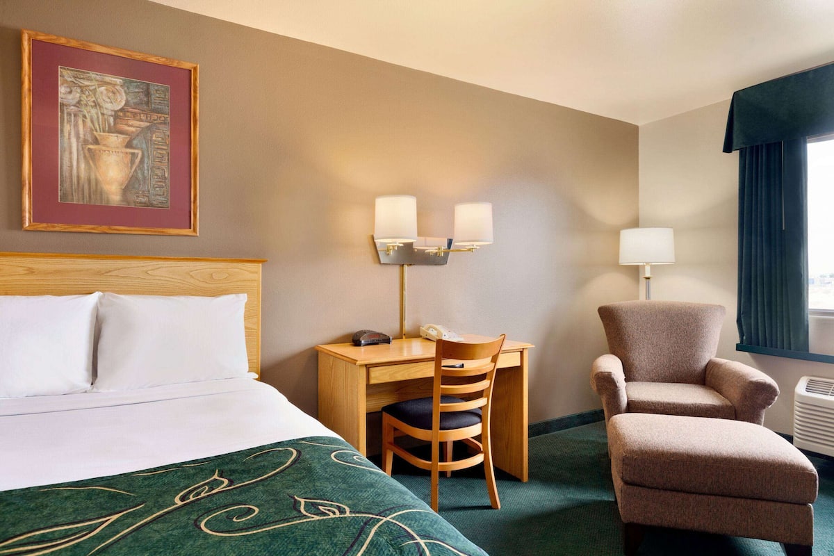 A hotel room with a bed, a desk with a chair, a lamp, and a window with curtains.