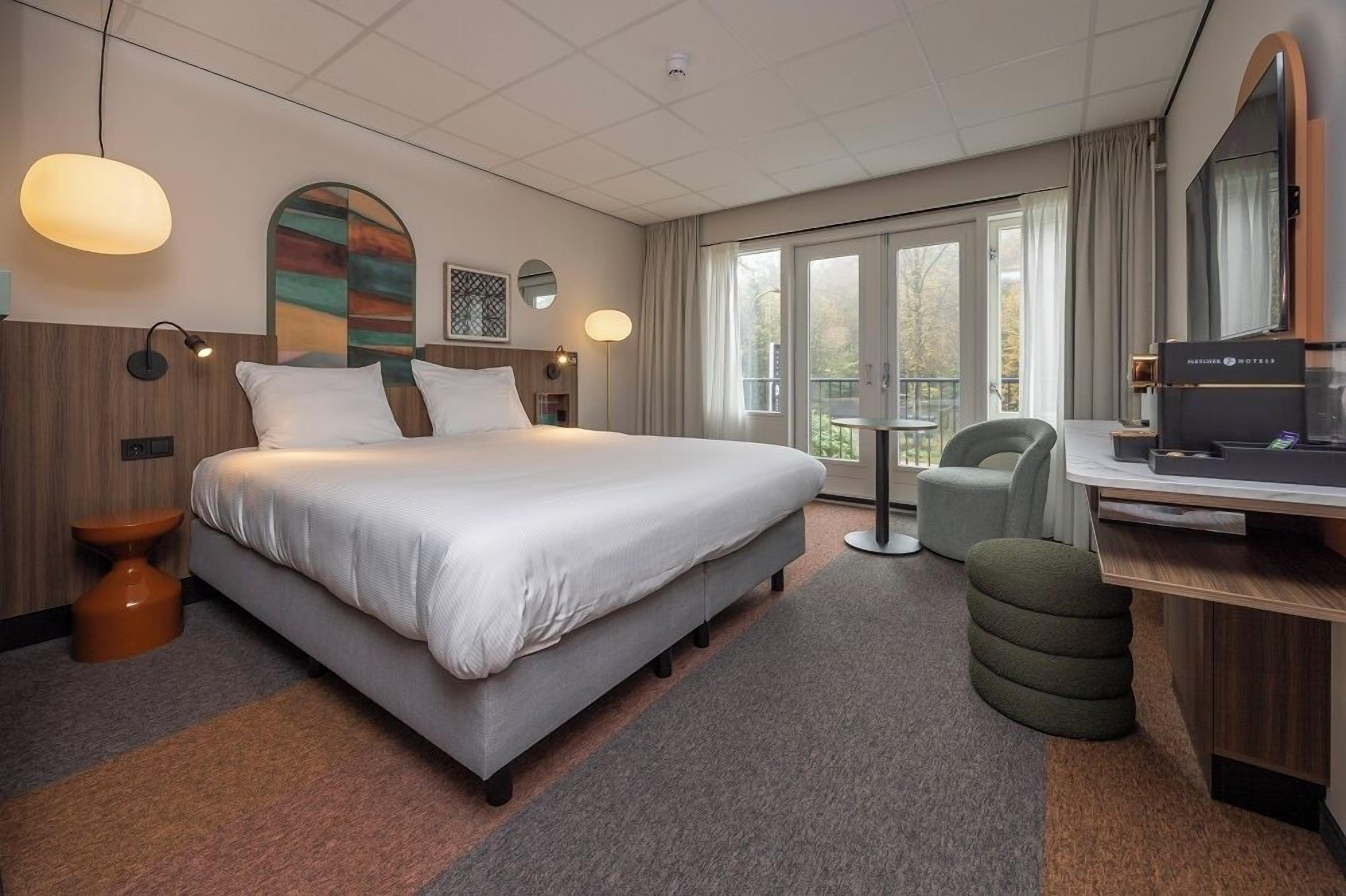 A hotel room with a large bed, a desk, a chair, a TV, and a balcony with a view.