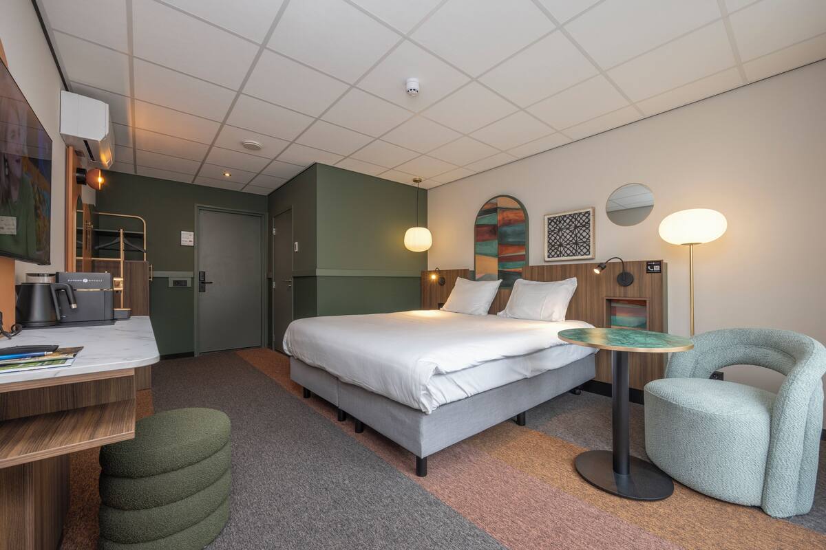 A modern hotel room with a bed, desk, chair, and a small table.