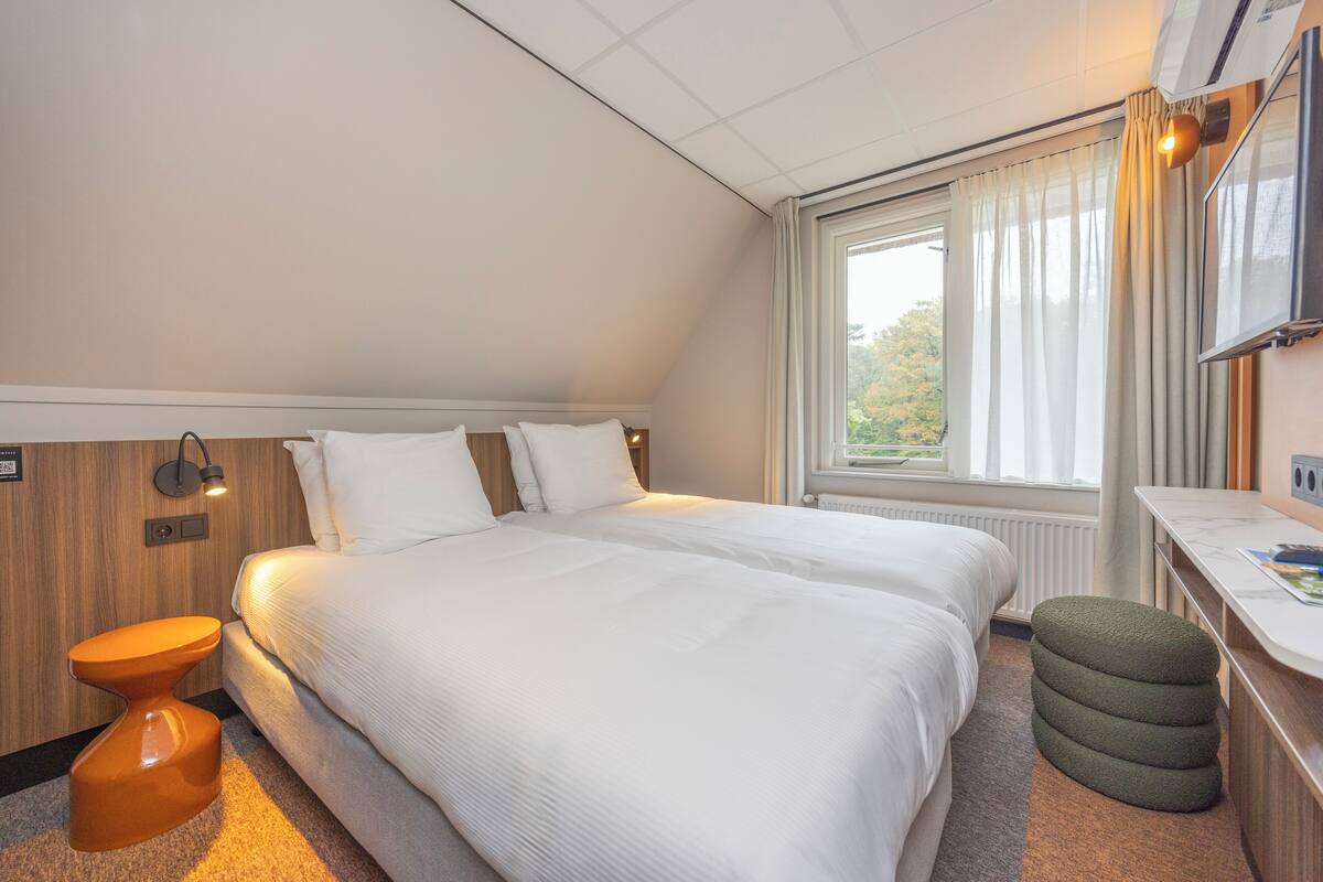A hotel room with a large bed, a small stool, a desk, and a window with curtains.