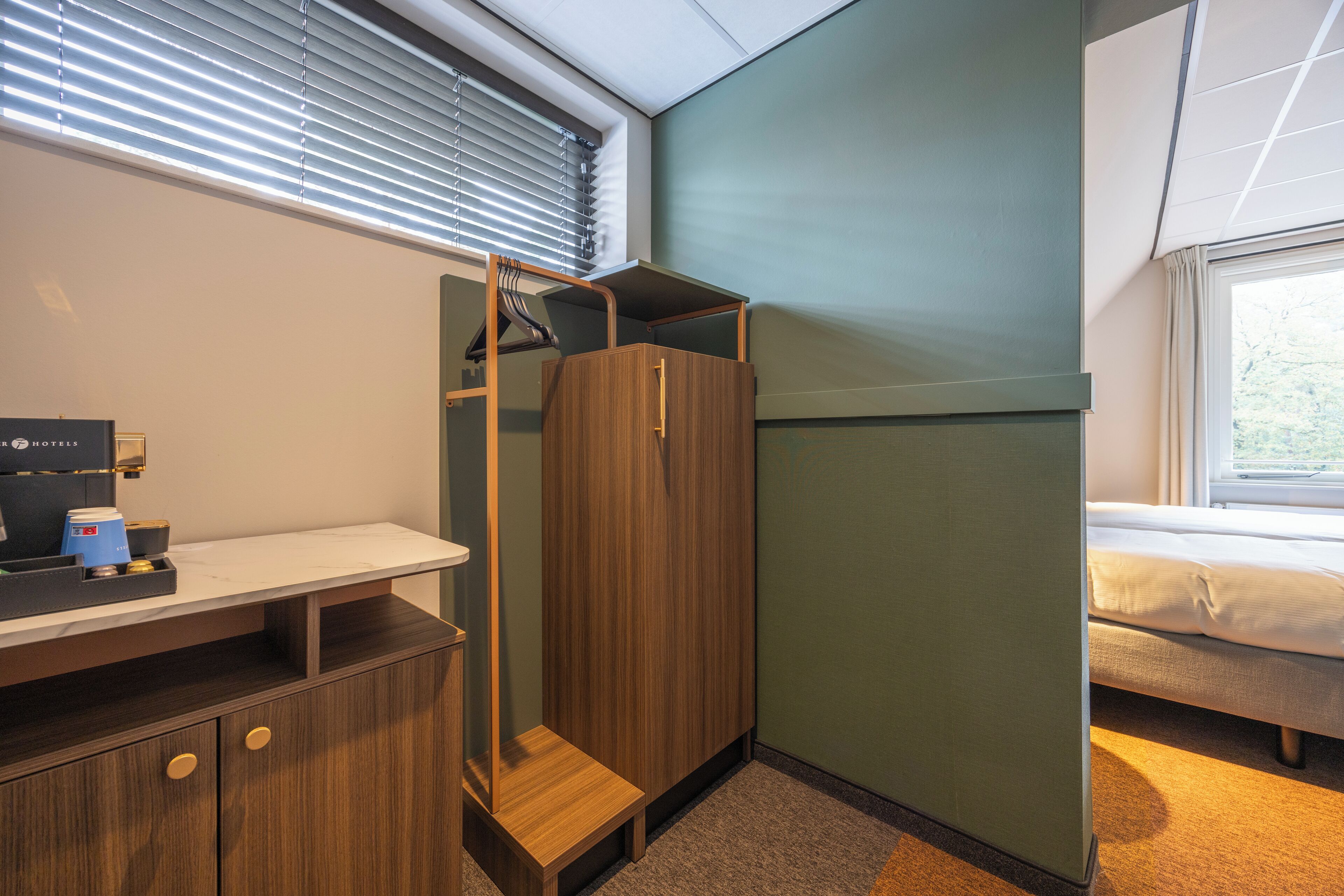 Een kleine hotelkamer met een bed, een bureau met koffiezetapparaat, een kledingkast en een raam met jaloezieën.
