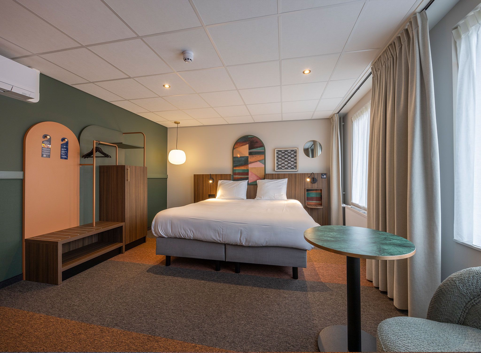 A hotel room with a bed, bedside table, wardrobe, and a view of the outdoors.
