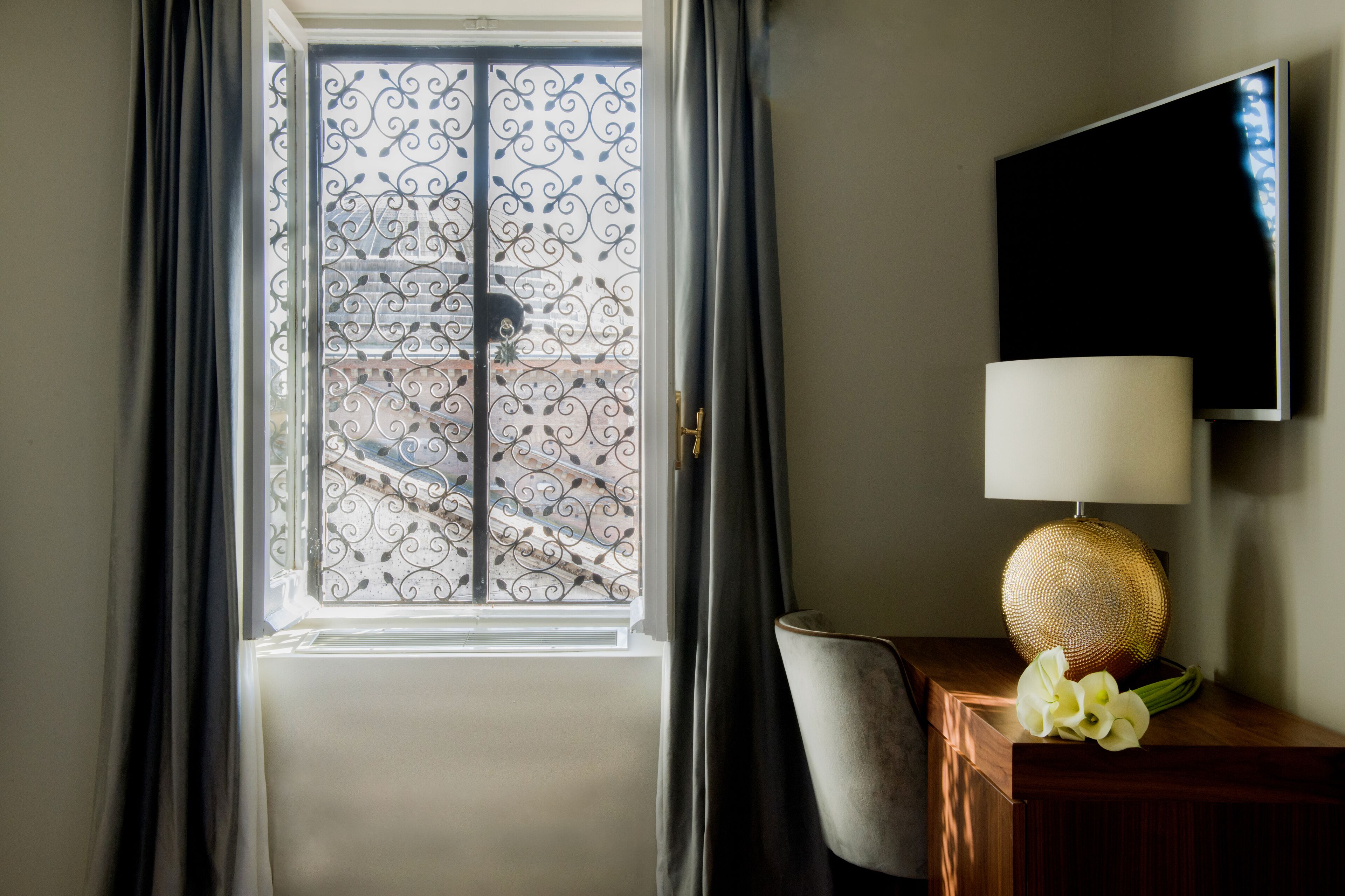 A room with a window featuring decorative ironwork, a flat-screen TV mounted on the wall, a lamp with a textured shade, and a chair.