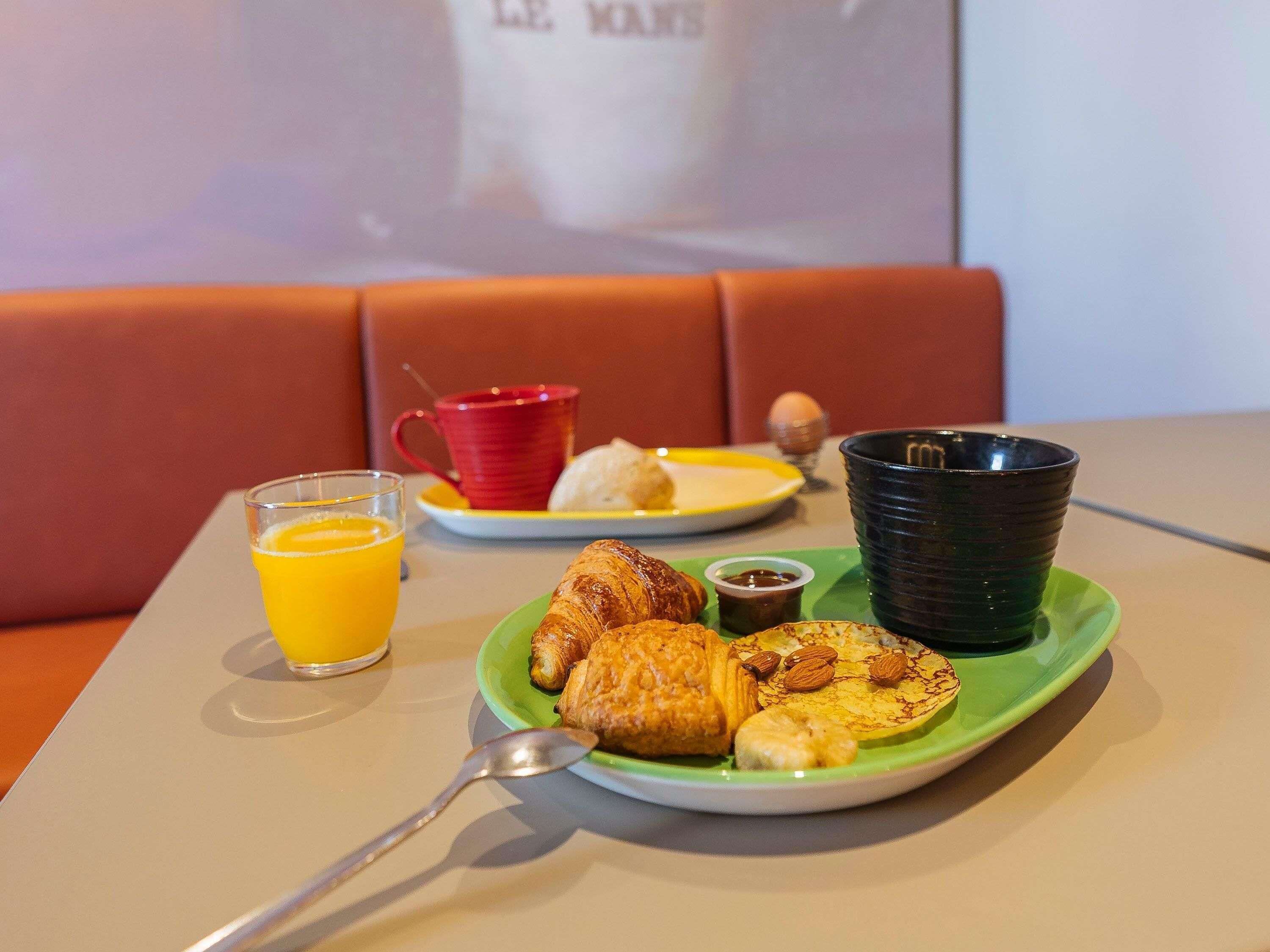 A breakfast setting with a plate of croissants, a glass of orange juice, a red cup, and a black cup on a table.