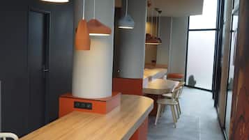 Ein modernes Interieur mit einem langen Holztisch und blauen Stühlen, Pendelleuchten an der Decke und Blick durch große Fenster nach draußen.
