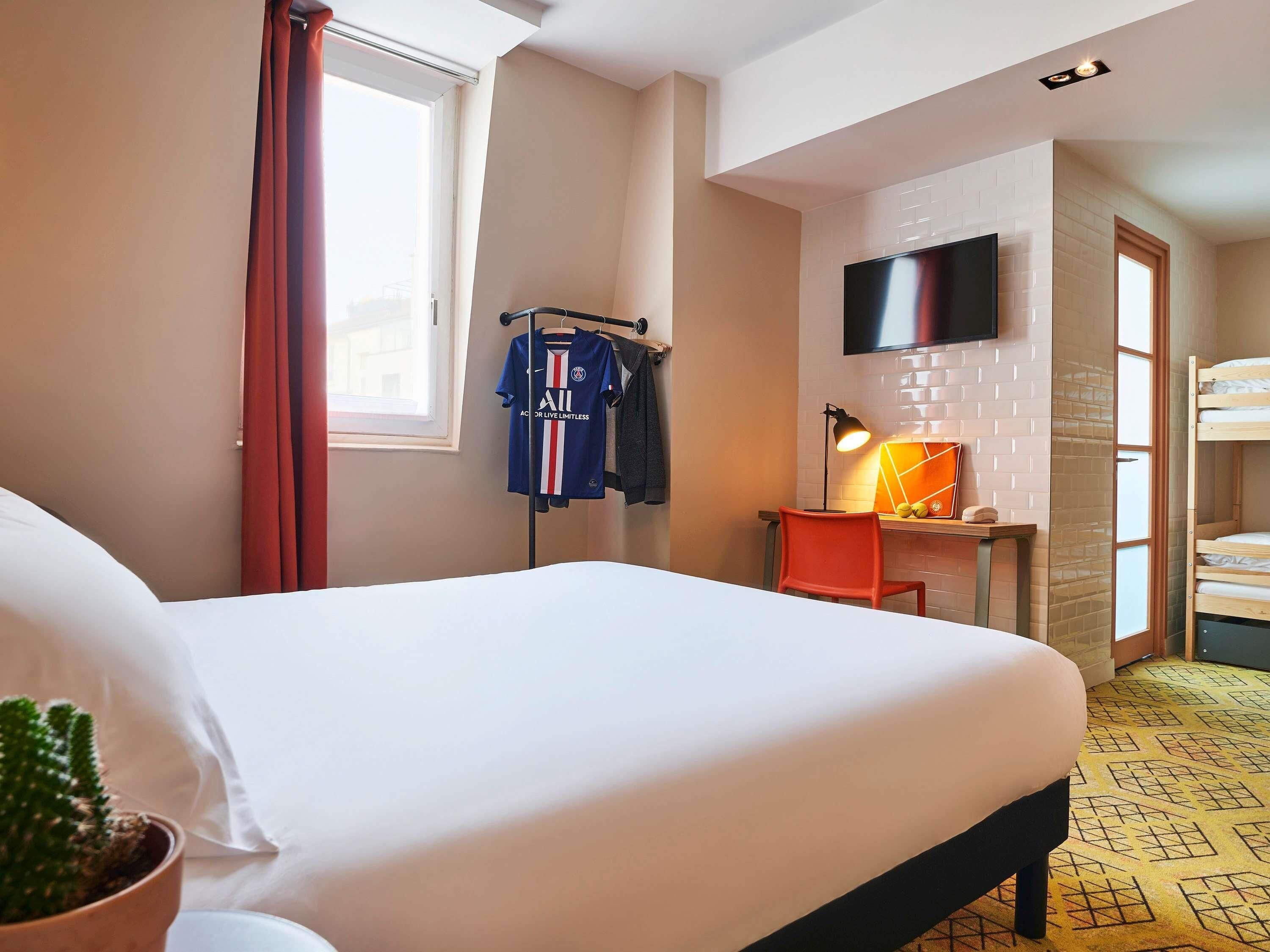 Une chambre d’hôtel avec un lit, un bureau et une chaise, un téléviseur fixé au mur, une fenêtre avec des rideaux et, au fond, un lit superposé.
