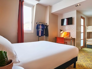 Une chambre d’hôtel avec un lit, un bureau et une chaise, un téléviseur fixé au mur, une fenêtre avec des rideaux et, au fond, un lit superposé.