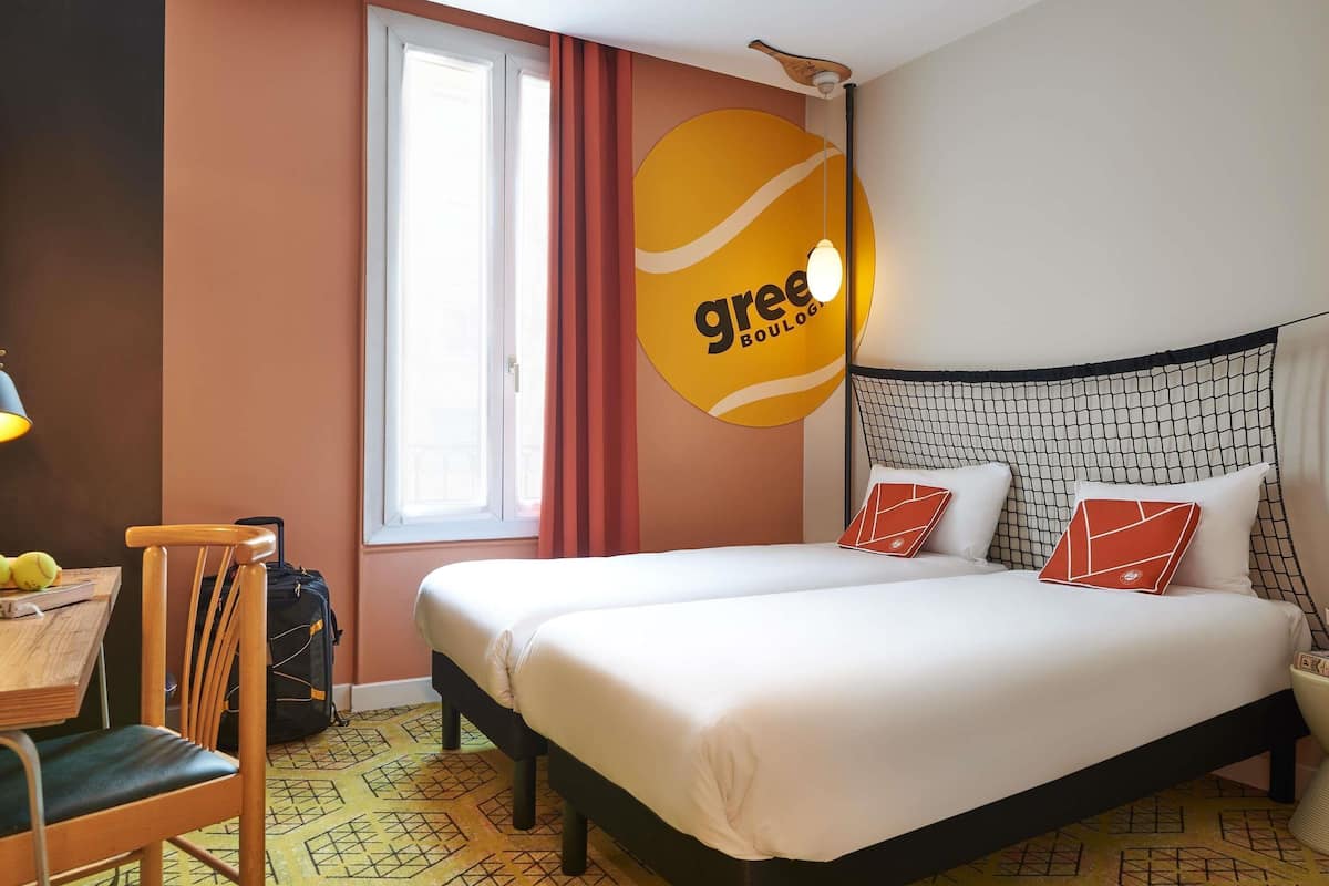 Une chambre dâhĂŽtel avec un lit, un bureau, une chaise, une fenĂȘtre et un mur ornĂ© dâun logo.