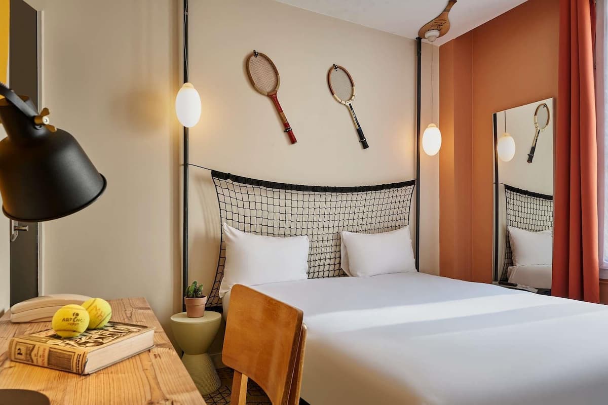 Une chambre dâhĂŽtel avec un lit, un bureau dotĂ© dâune lampe, une chaise, une petite table sur laquelle se trouvent un livre et des balles de tennis, et deux raquettes de tennis accrochĂ©es au mur.