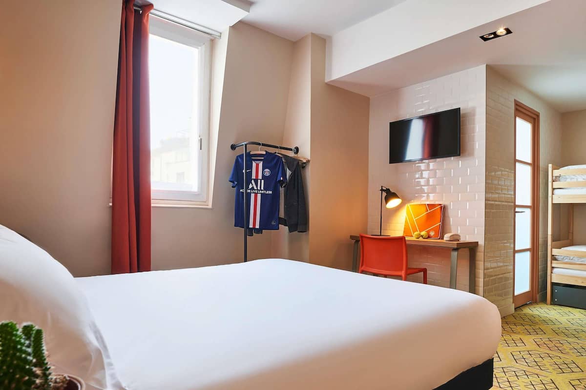 A hotel room with a bed, a desk with a chair, a TV mounted on the wall, a window with curtains, and a bunk bed in the background.