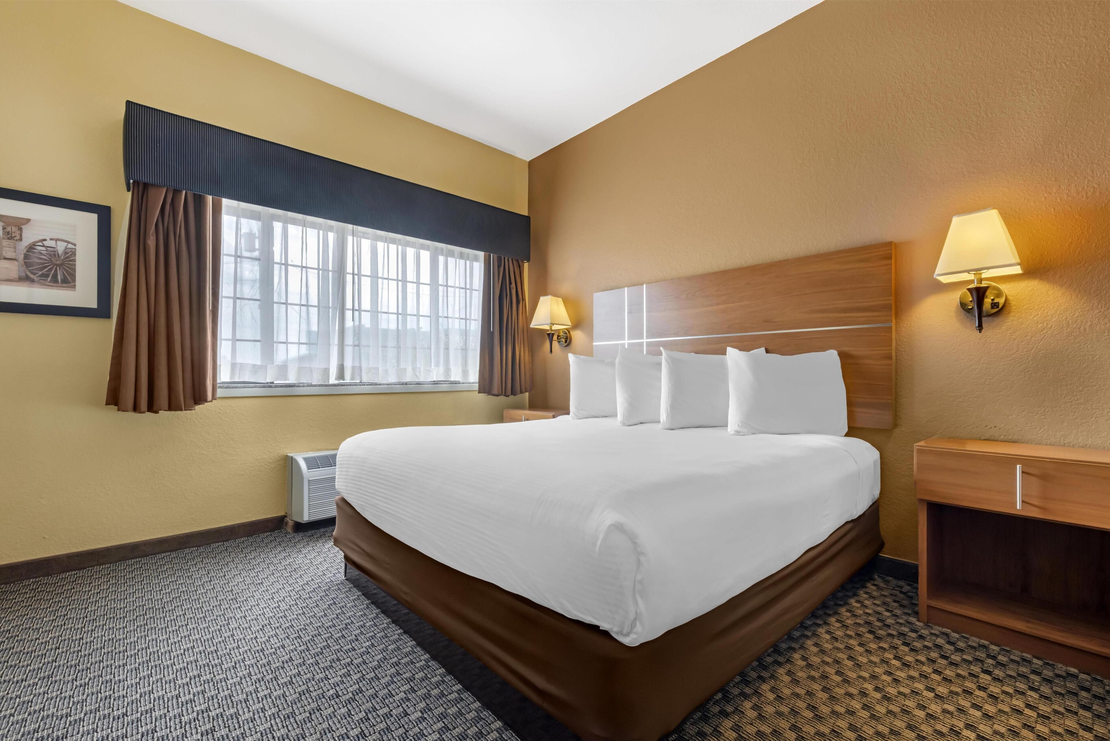 A hotel room with a large bed, two bedside lamps, a window with curtains, and a framed picture on the wall.