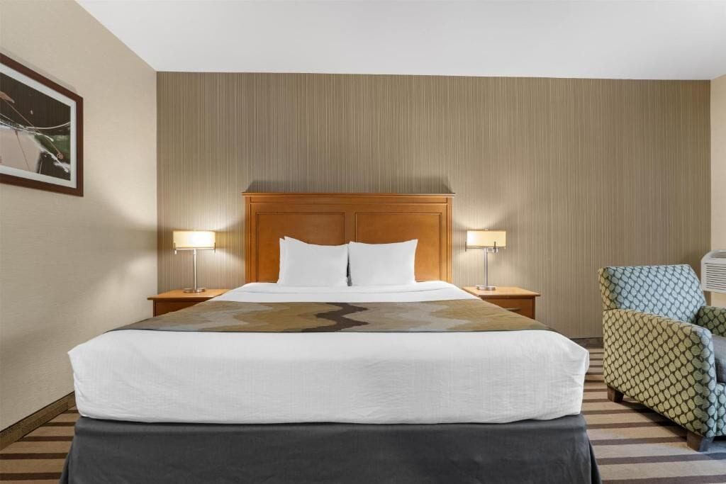 A hotel room with a large bed, two bedside tables with lamps, a patterned armchair, and a wall-mounted air conditioning unit.