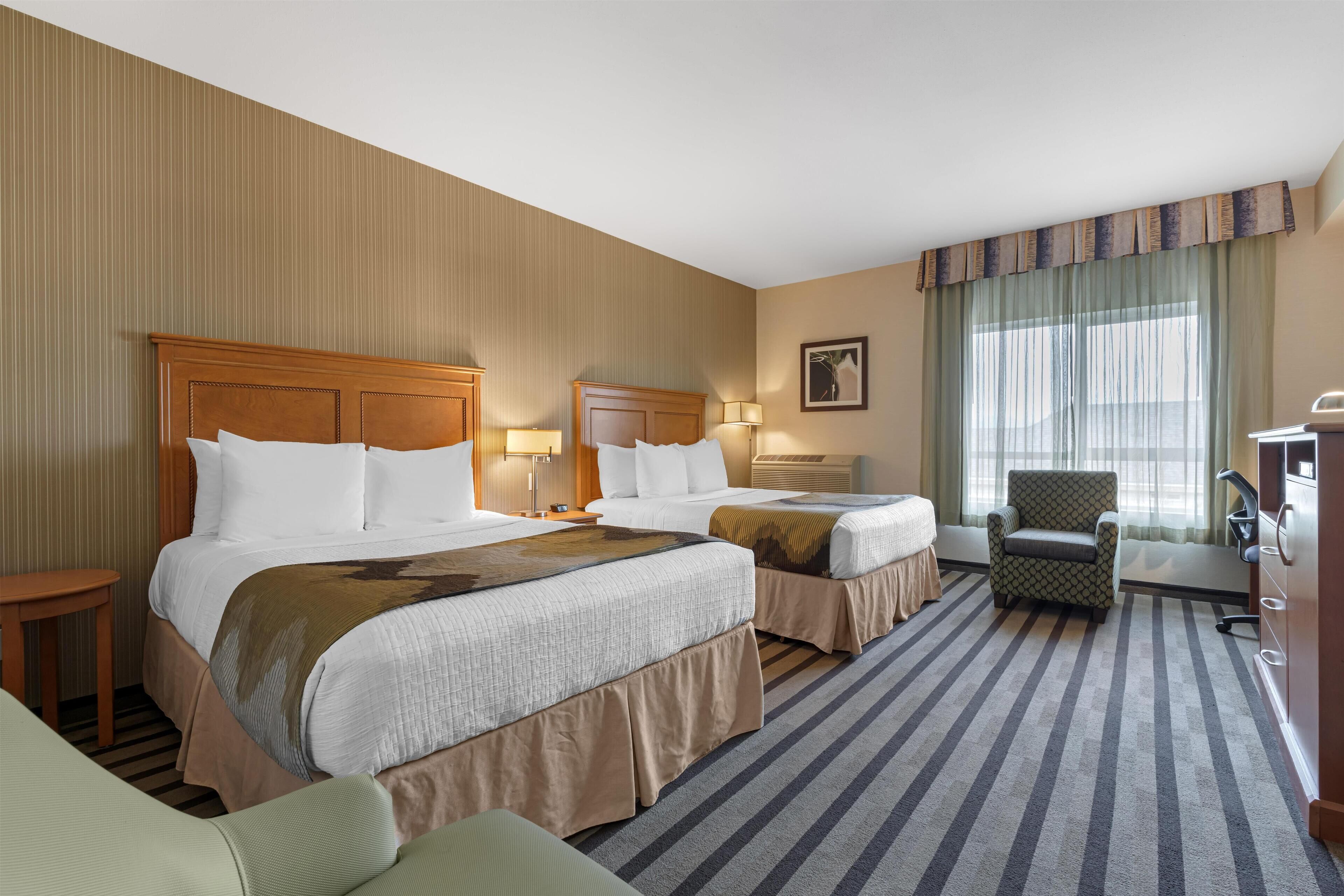 A hotel room with two beds, a chair, a desk, and a large window with curtains.