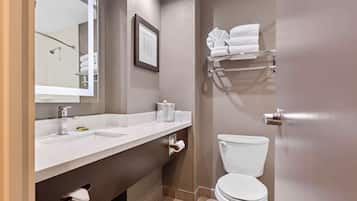 A modern bathroom with a white sink, mirror, and toilet. There is a towel rack above the toilet and a framed picture on the wall.