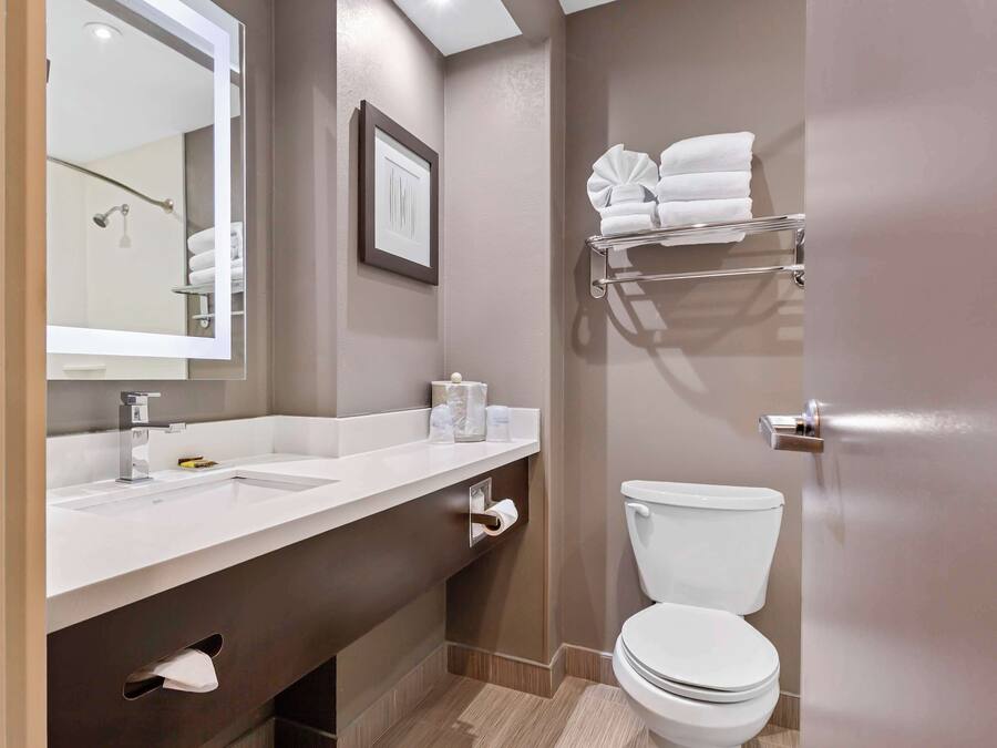Un baño moderno con un lavamanos, espejo y inodoro blancos. Hay un perchero sobre el inodoro y un cuadro en la pared.