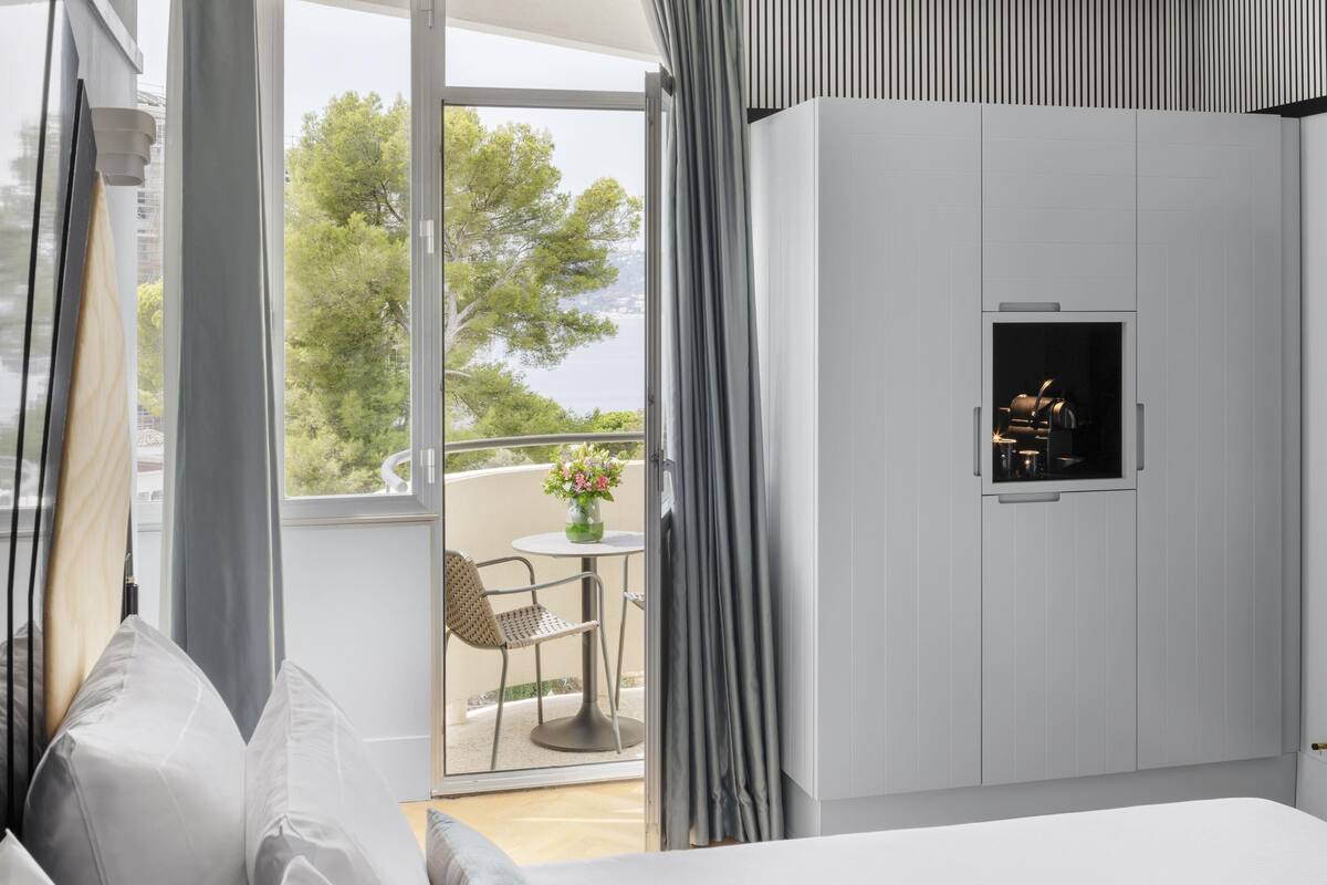 Ein Schlafzimmer mit Bett, Fernseher, einem Balkon mit Tisch und Stuhl sowie Blick auf Bäume und Gewässer.