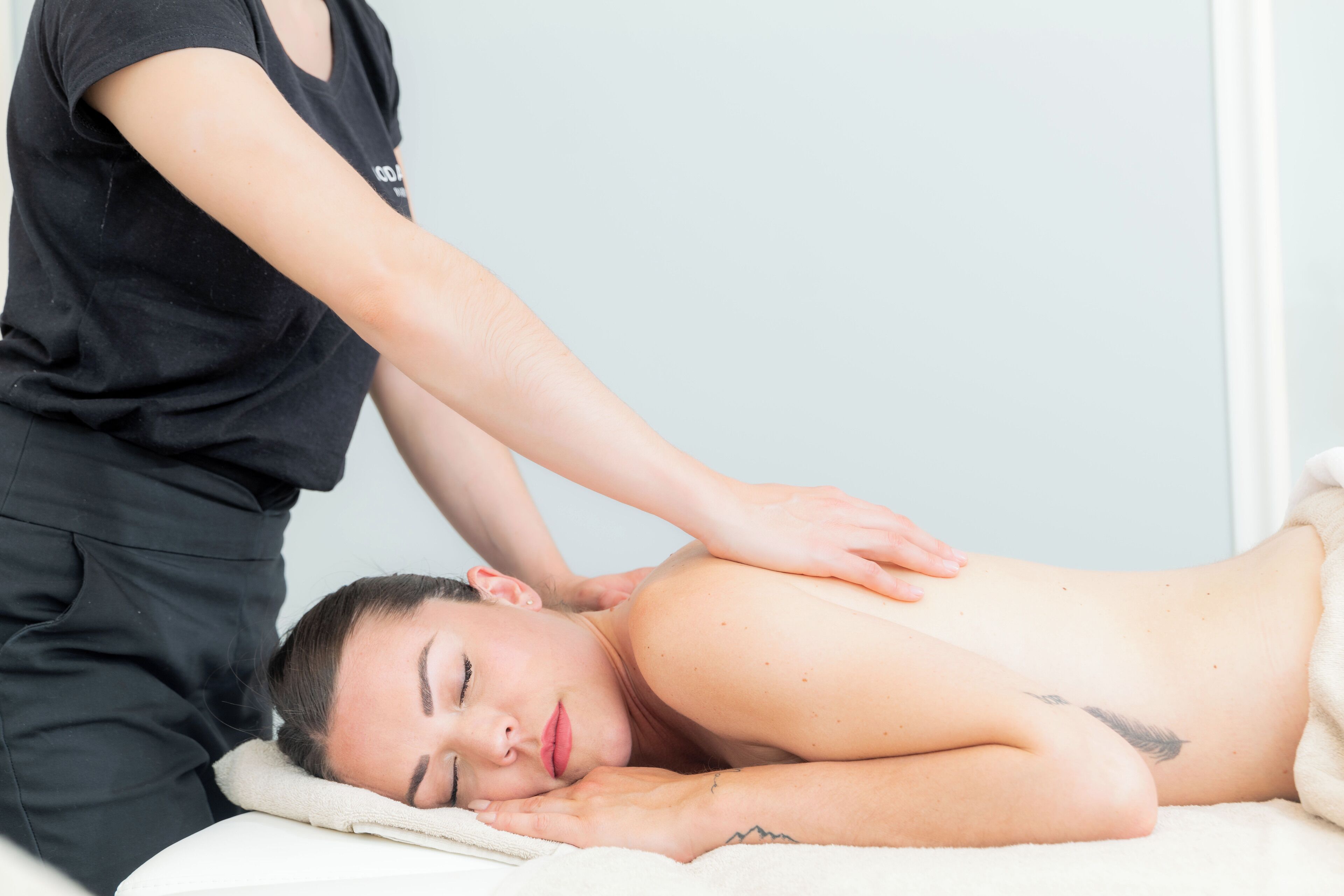 A person receiving a back massage in a room with a white bed and a plain wall.