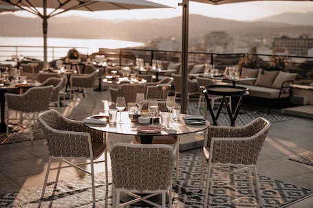 An outdoor dining area with round tables and wicker chairs, overlooking a body of water and cityscape.