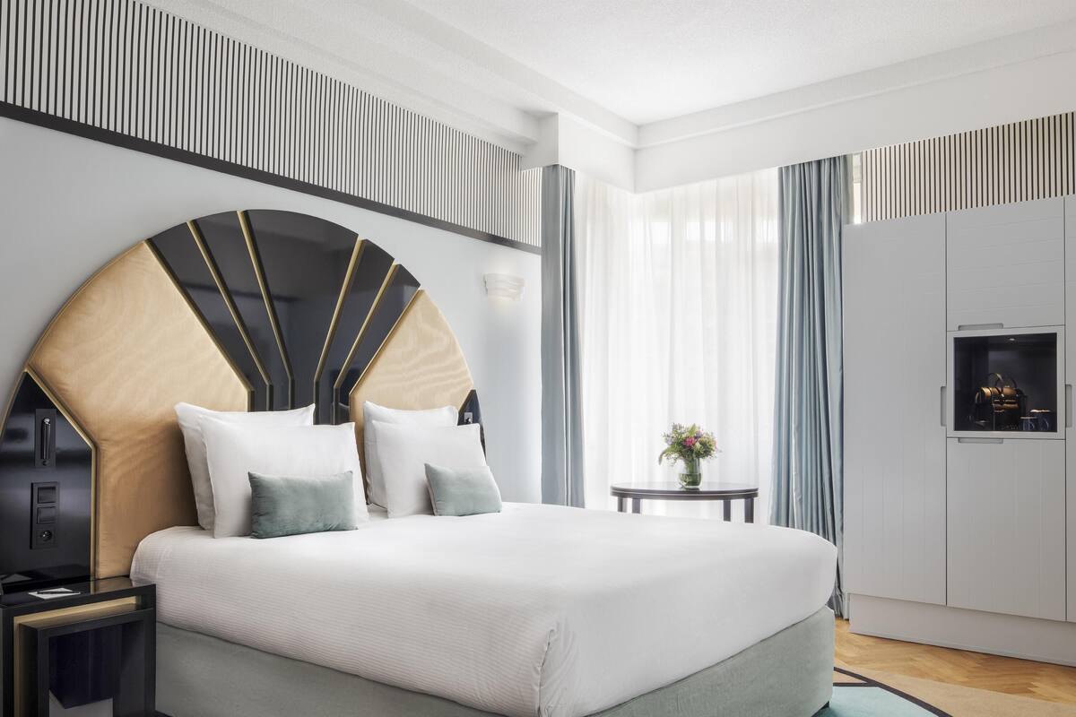 A modern hotel room with a large bed, a bedside table, a small round table with a vase, and a wardrobe with a mirror.