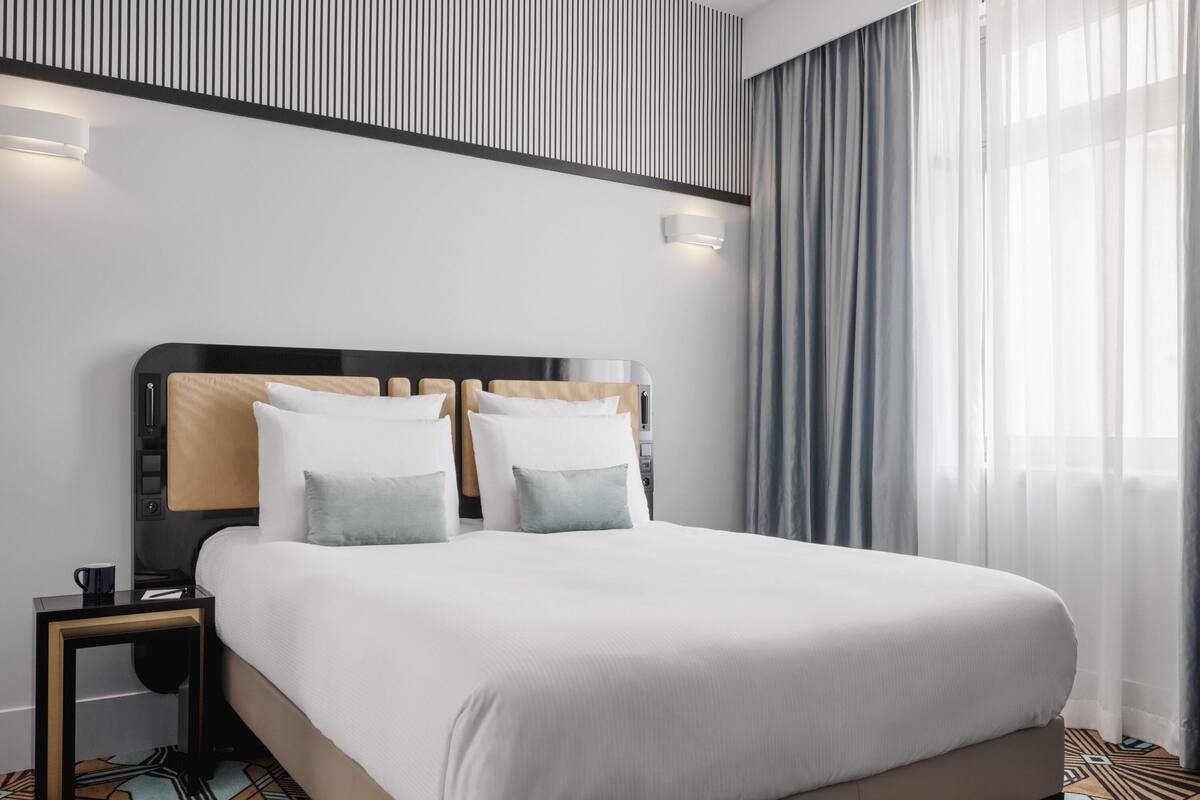 A modern hotel room with a large bed, a nightstand, a wall-mounted light, and a window with sheer curtains.