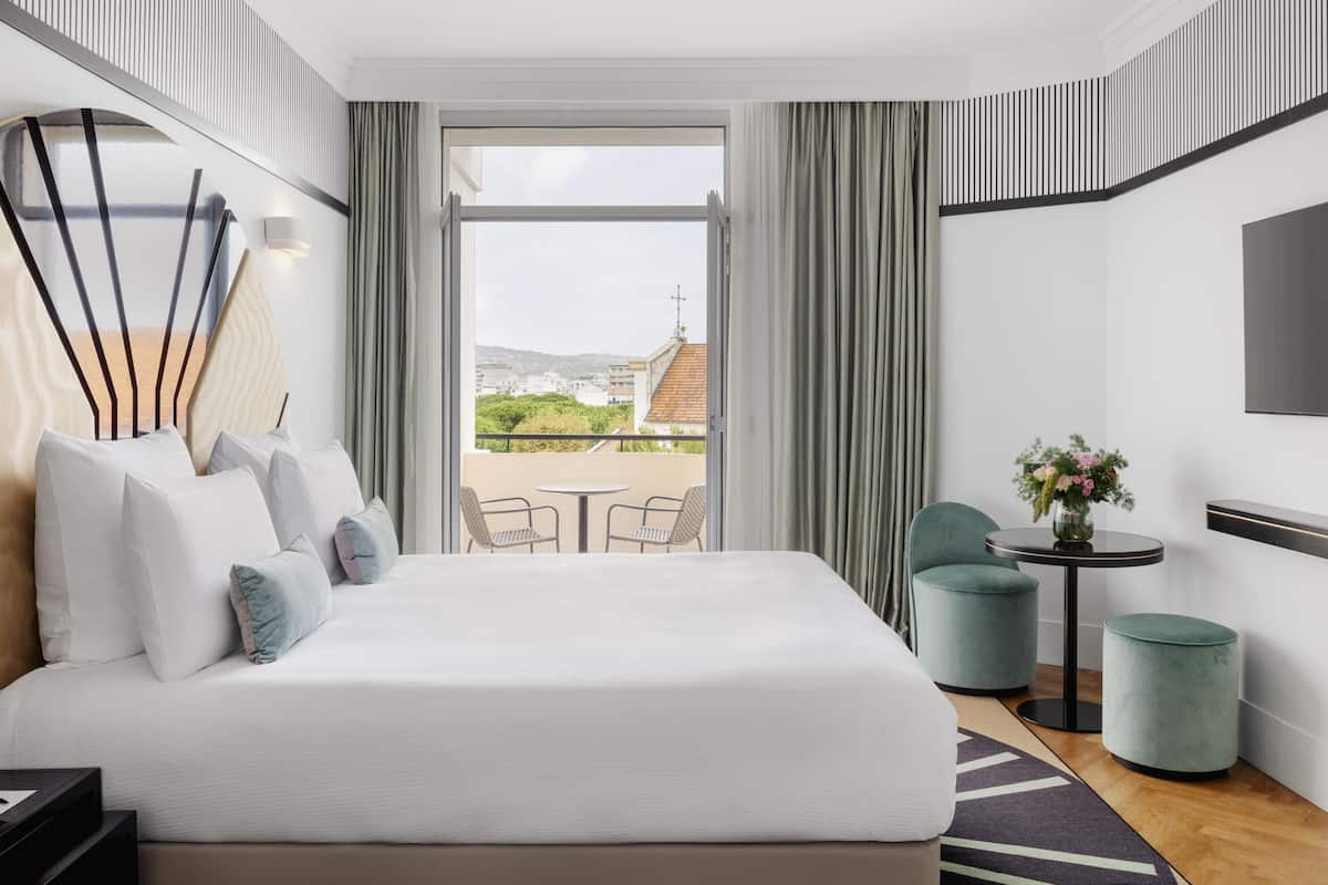 Une chambre d’hôtel moderne avec un grand lit, un balcon avec des chaises, une petite table agrémentée de fleurs et un téléviseur fixé au mur.